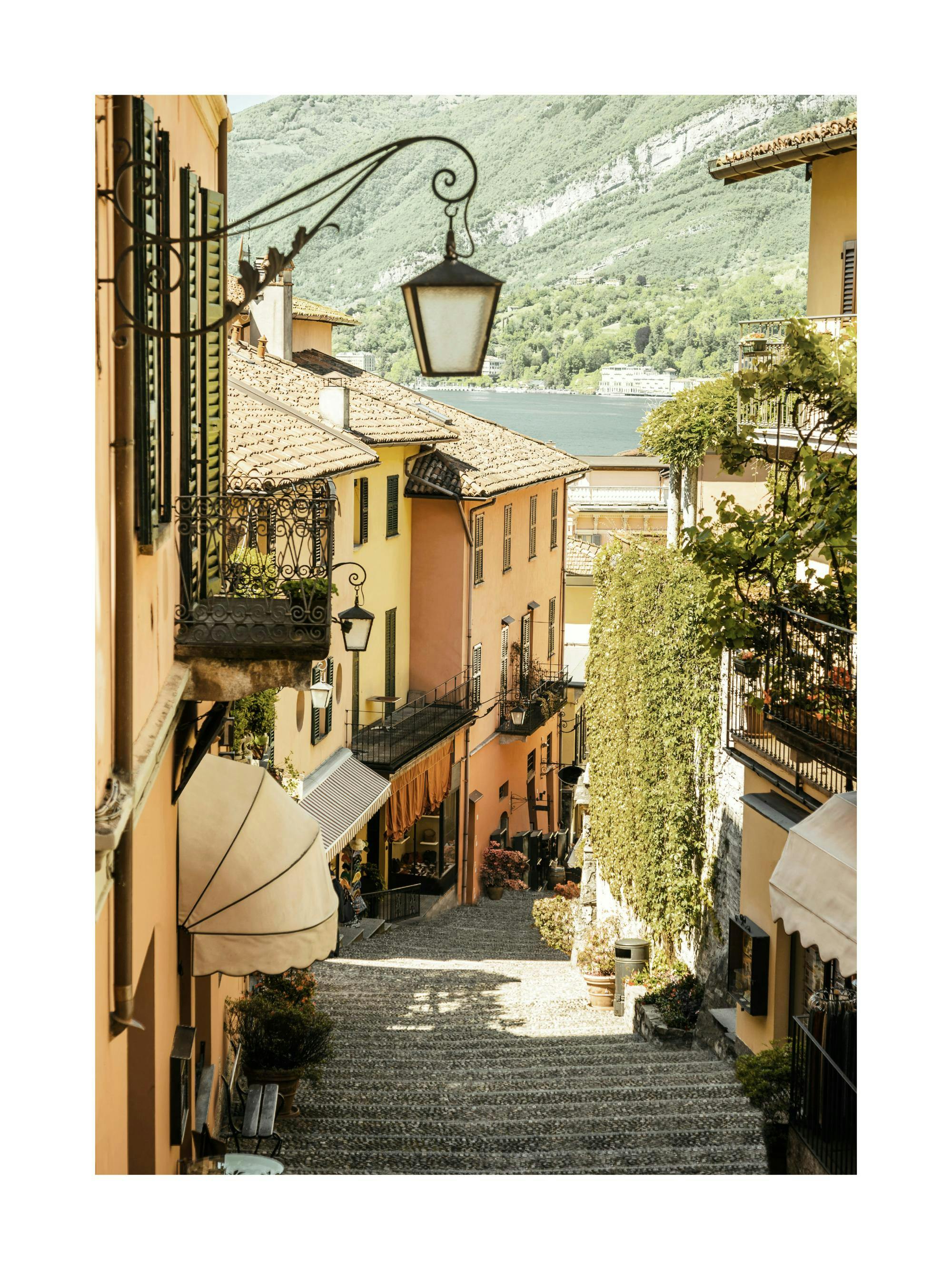Poster dune ruelle italienne en pente avec des bâtiments colorés, des balcons fleuris et un lac en arrière-plan.