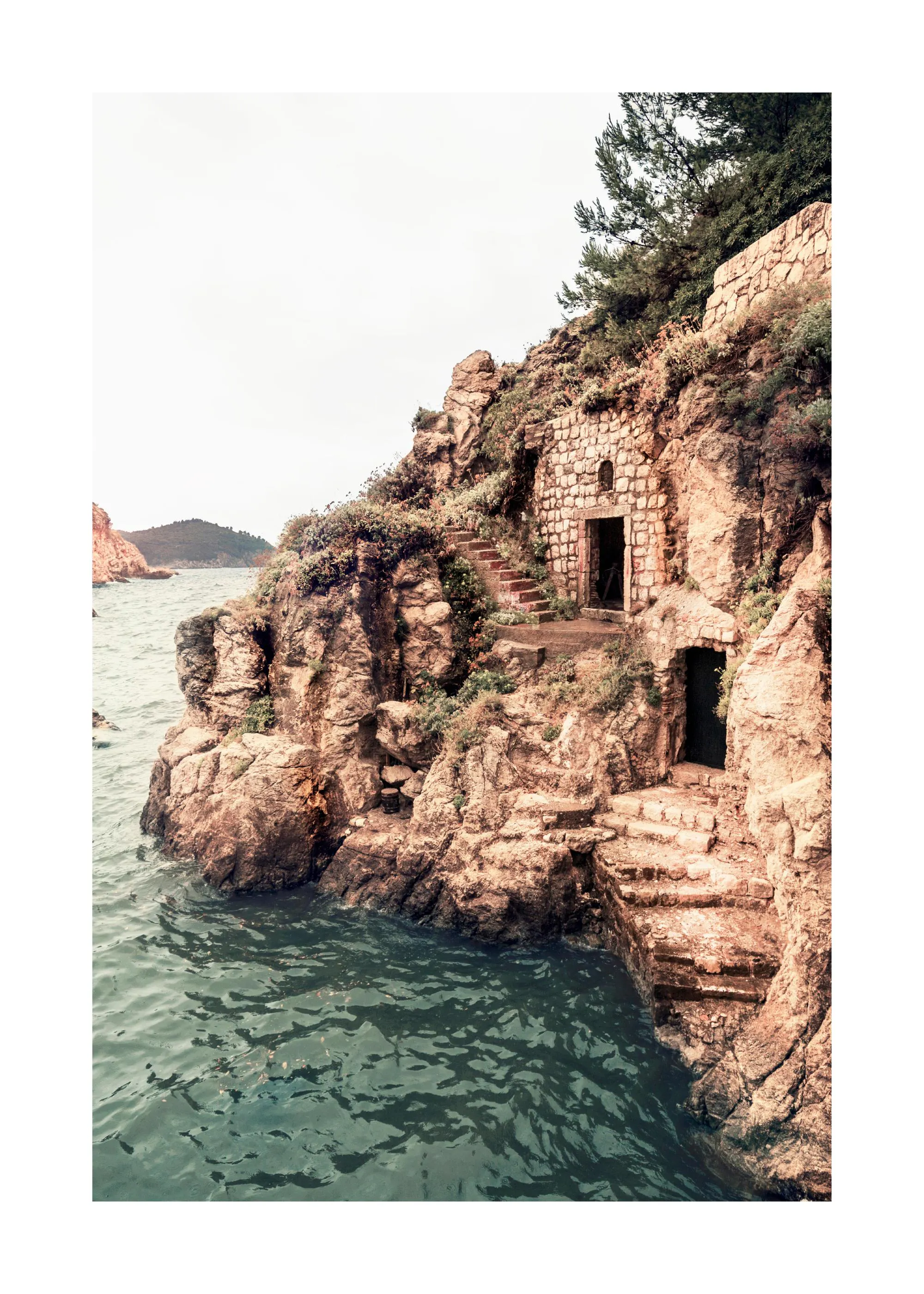 Poster einer felsigen Küste mit Steintreppen und Höhlen am Meer unter hellem Himmel.