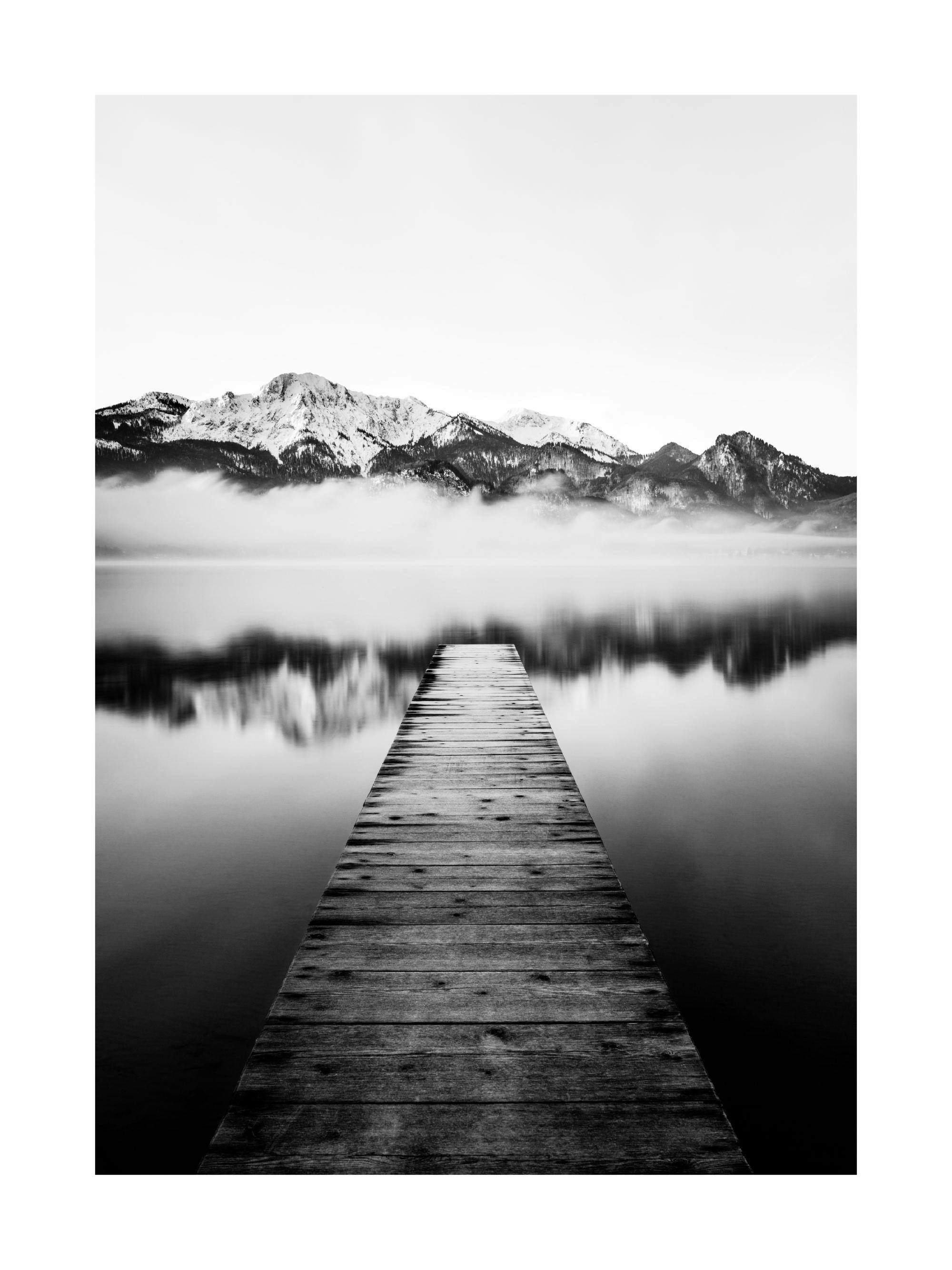 Czarno-biały plakat przedstawiający długie, drewniane molo na spokojnym jeziorze z górskim krajobrazem w tle.