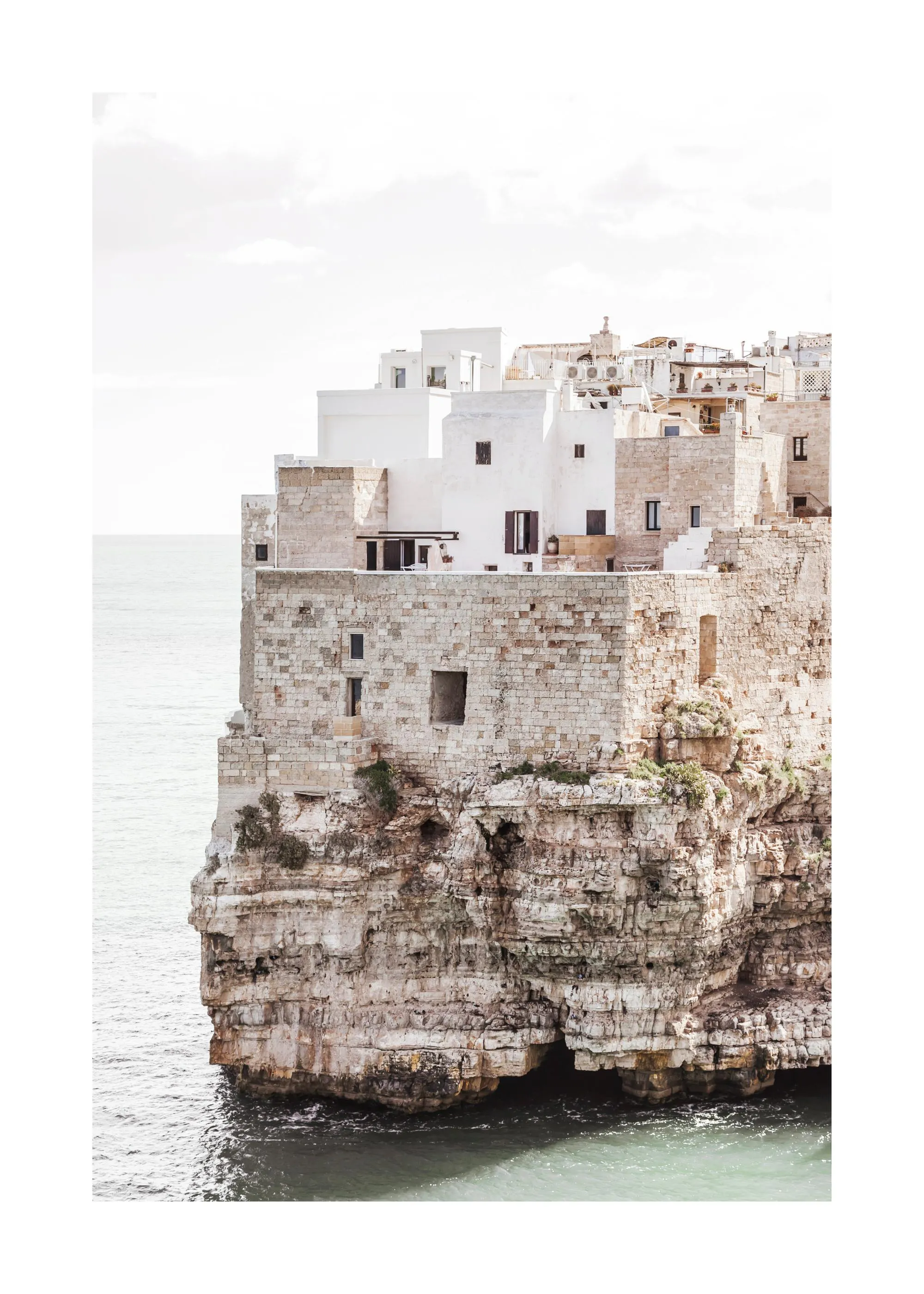 Poster: Polignano a Mare, weiße Häuser auf hohen Felsen direkt am Meer unter hellem Himmel.