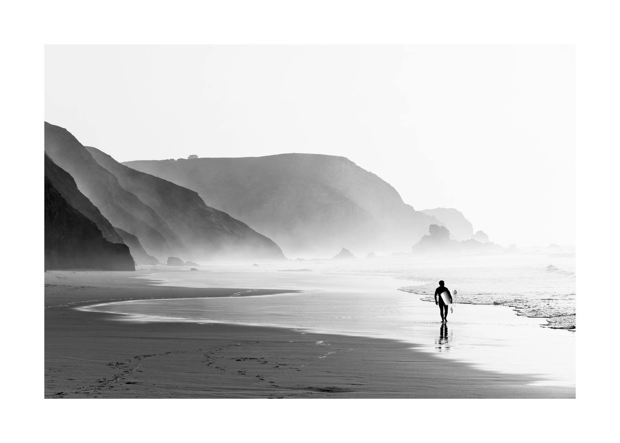 Un poster en noir et blanc dun surfeur marchant sur la plage avec des falaises et du brouillard à larrière-plan.