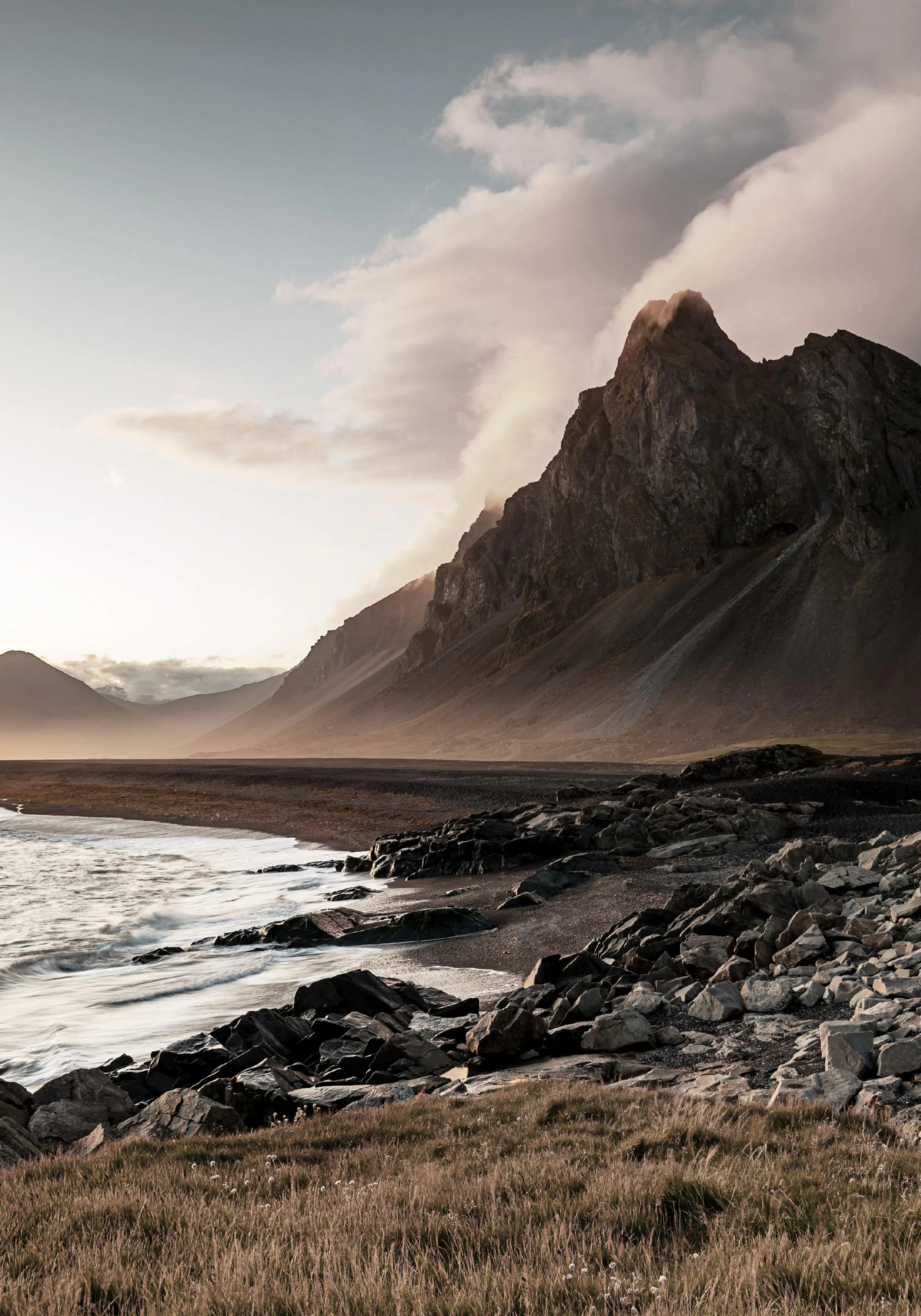 A poster featuring a rugged mountain range along a dark rocky coastline with waves gently washing ashore, under a soft, cloudy s