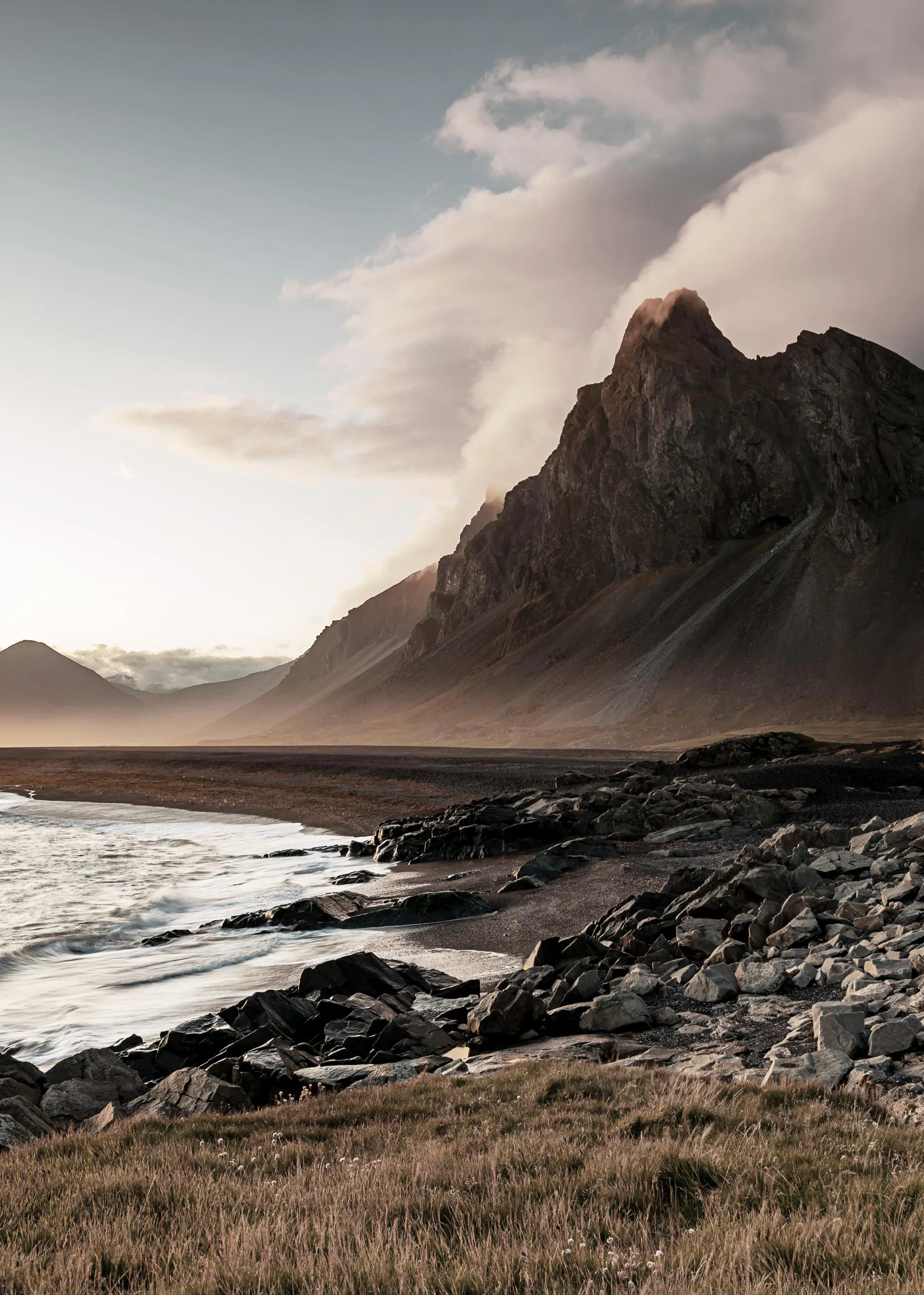 Ein Poster einer rauen Küstenlandschaft mit dunklen Felsen, Wellen und einem majestätischen Berg unter einem bewölkten Himmel.