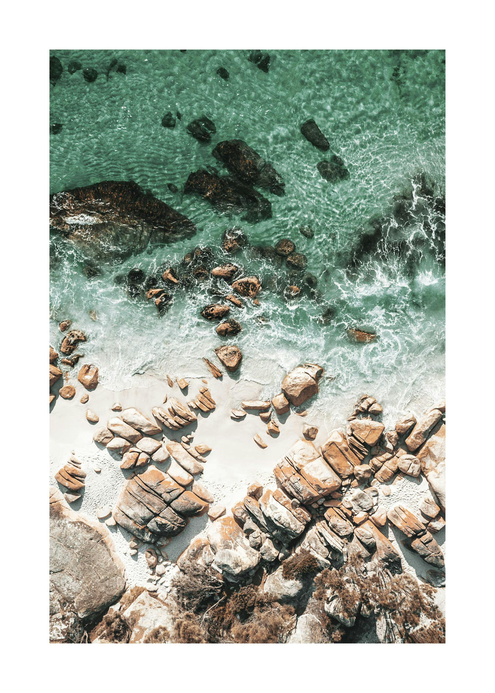 A poster depicting an aerial view of a vibrant turquoise ocean with crashing waves next to a sandy beach with large rocks.