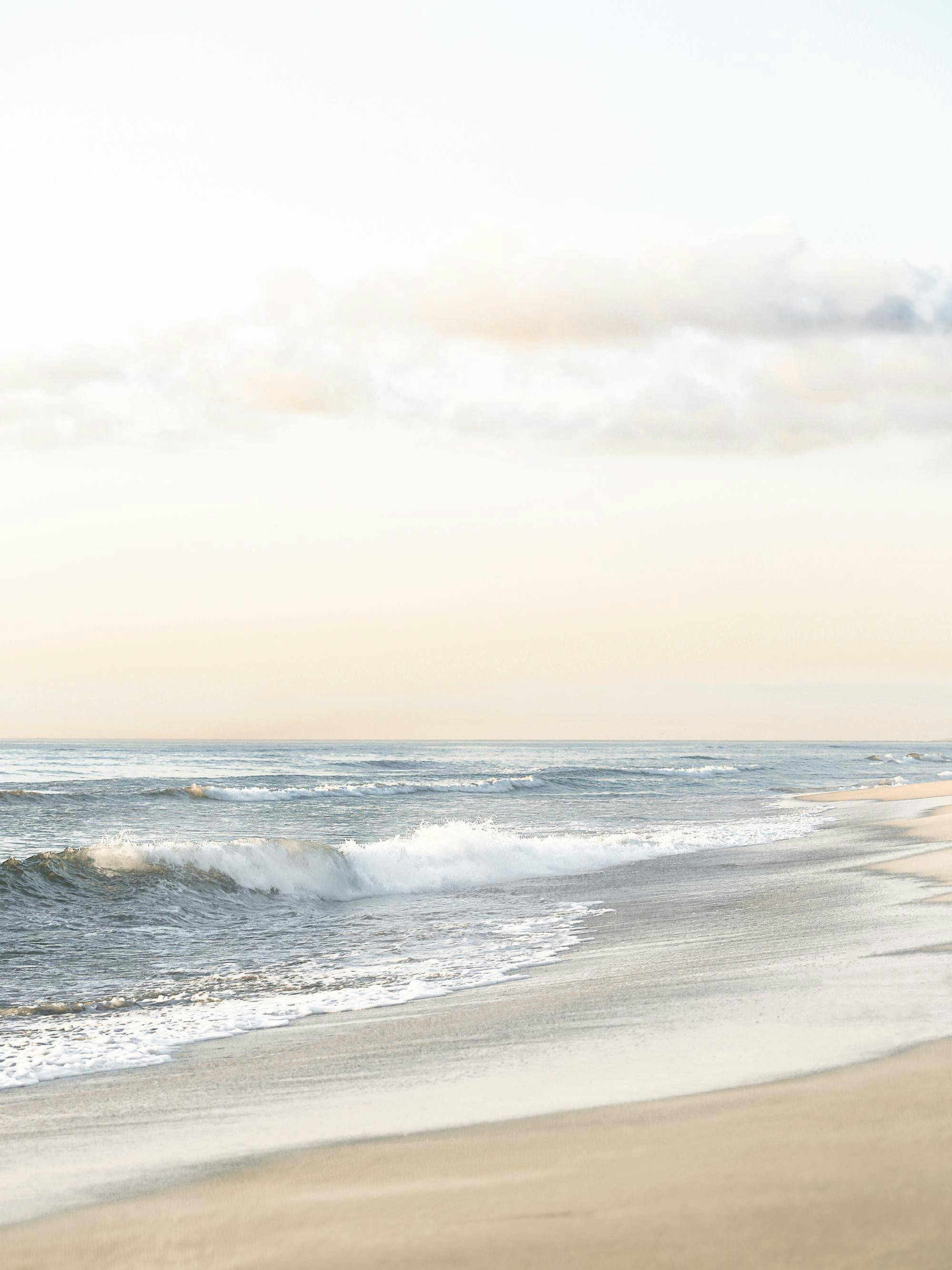 Poster cu o plajă vastă, cu valuri spumoase, nisip auriu și cer deschis, într-o lumină blândă.