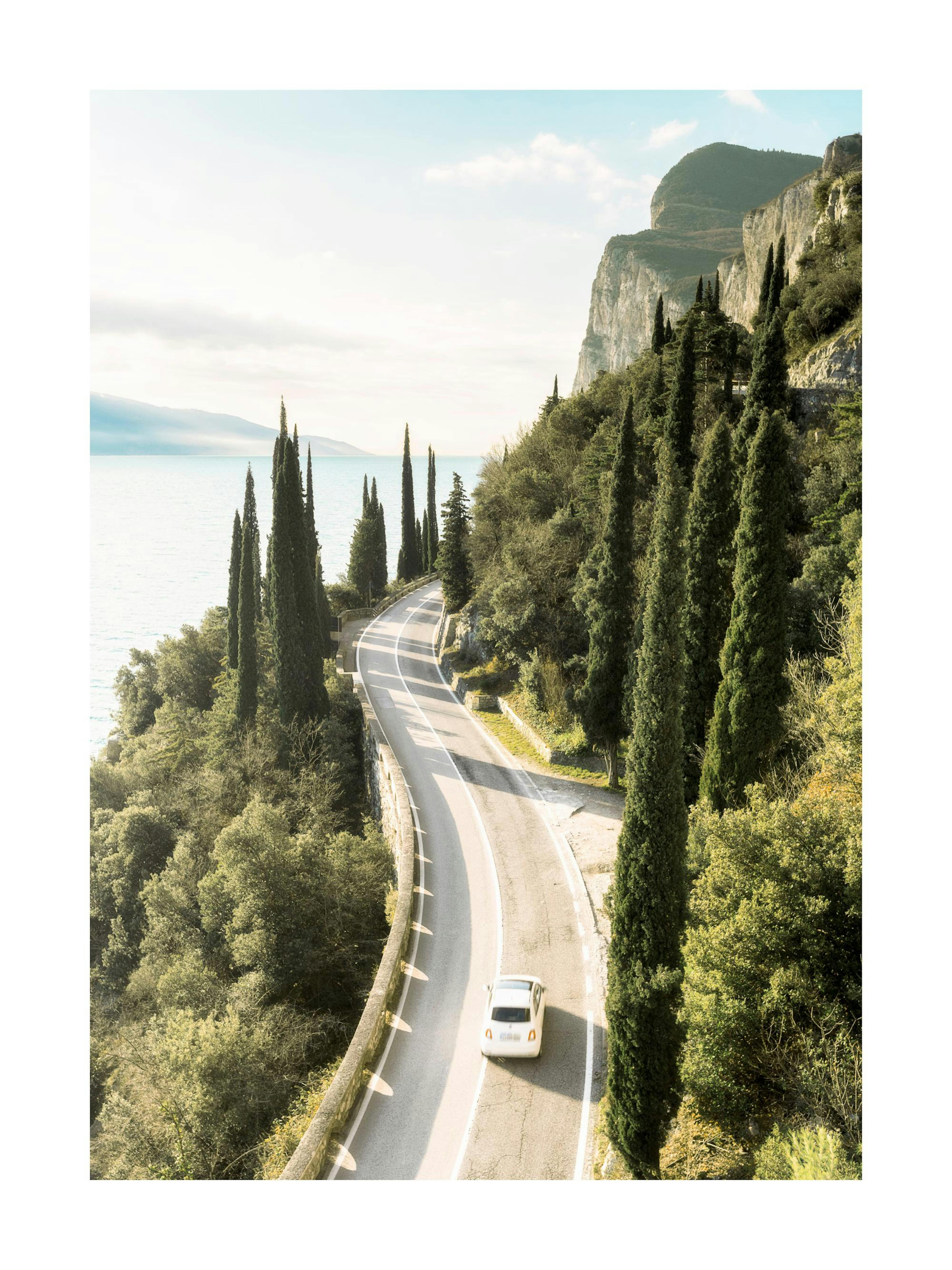 Póster de un coche blanco en una carretera serpenteante junto a un lago y montañas, con cipreses verdes.