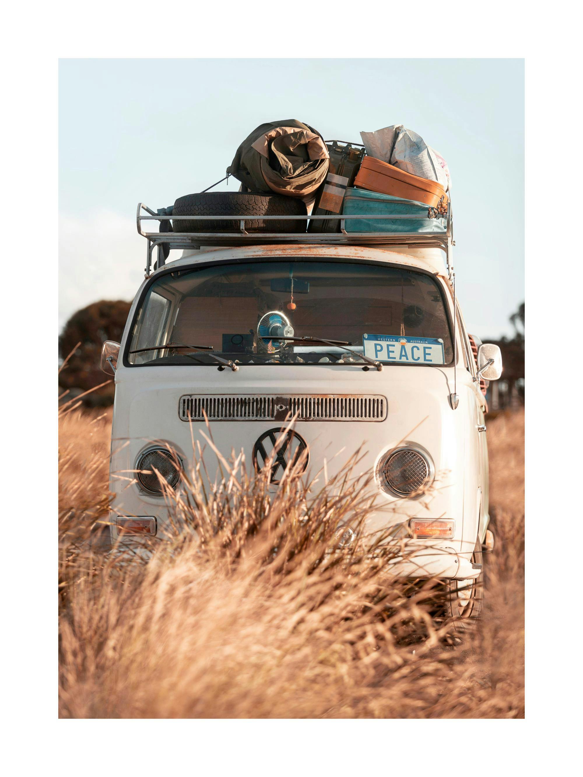 Plakat przedstawiający stary, biały van kempingowy z bagażem na dachu, stojący w wysokiej trawie.