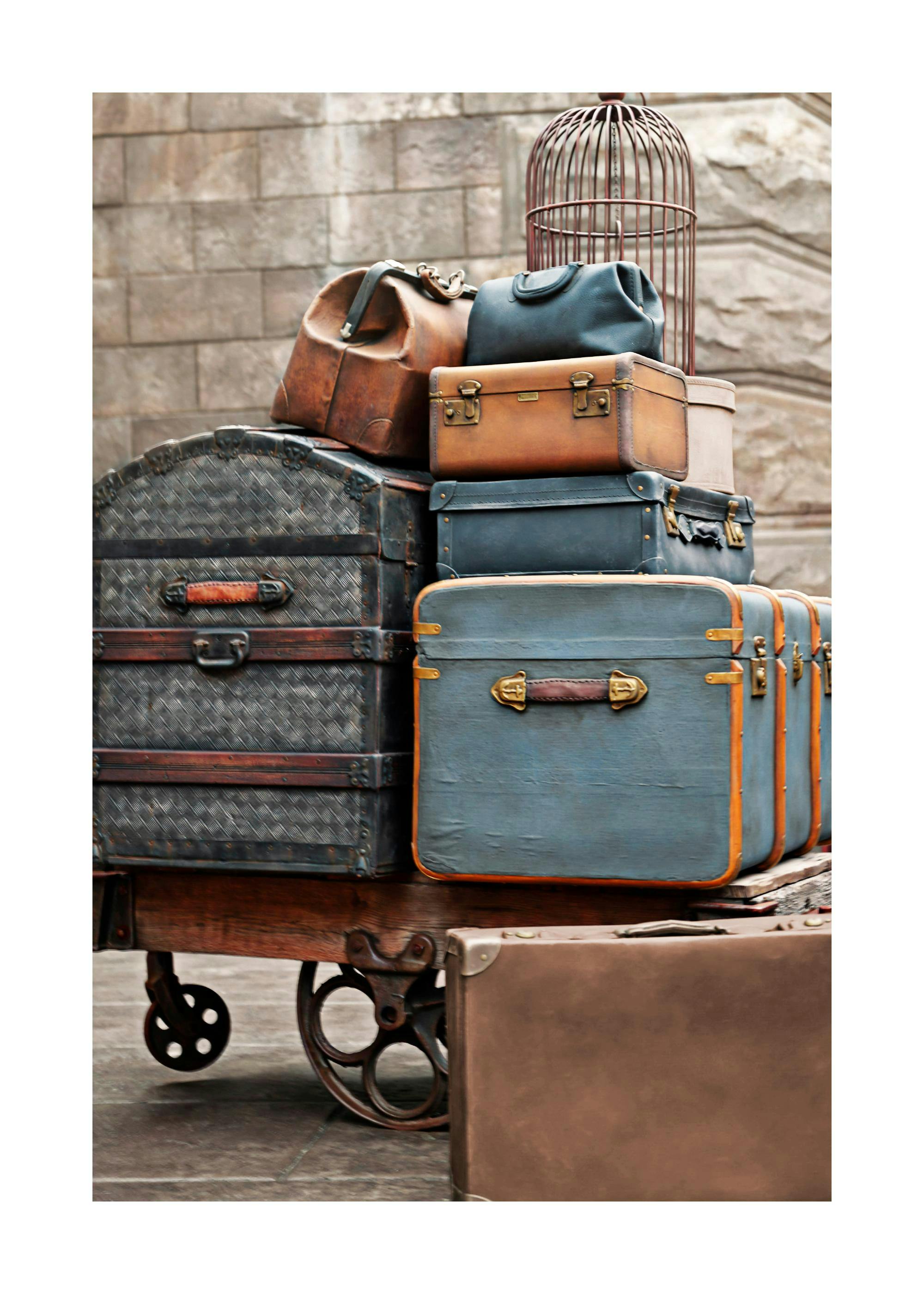 A poster featuring a stack of vintage suitcases and bags on a wooden cart, with a birdcage on top.