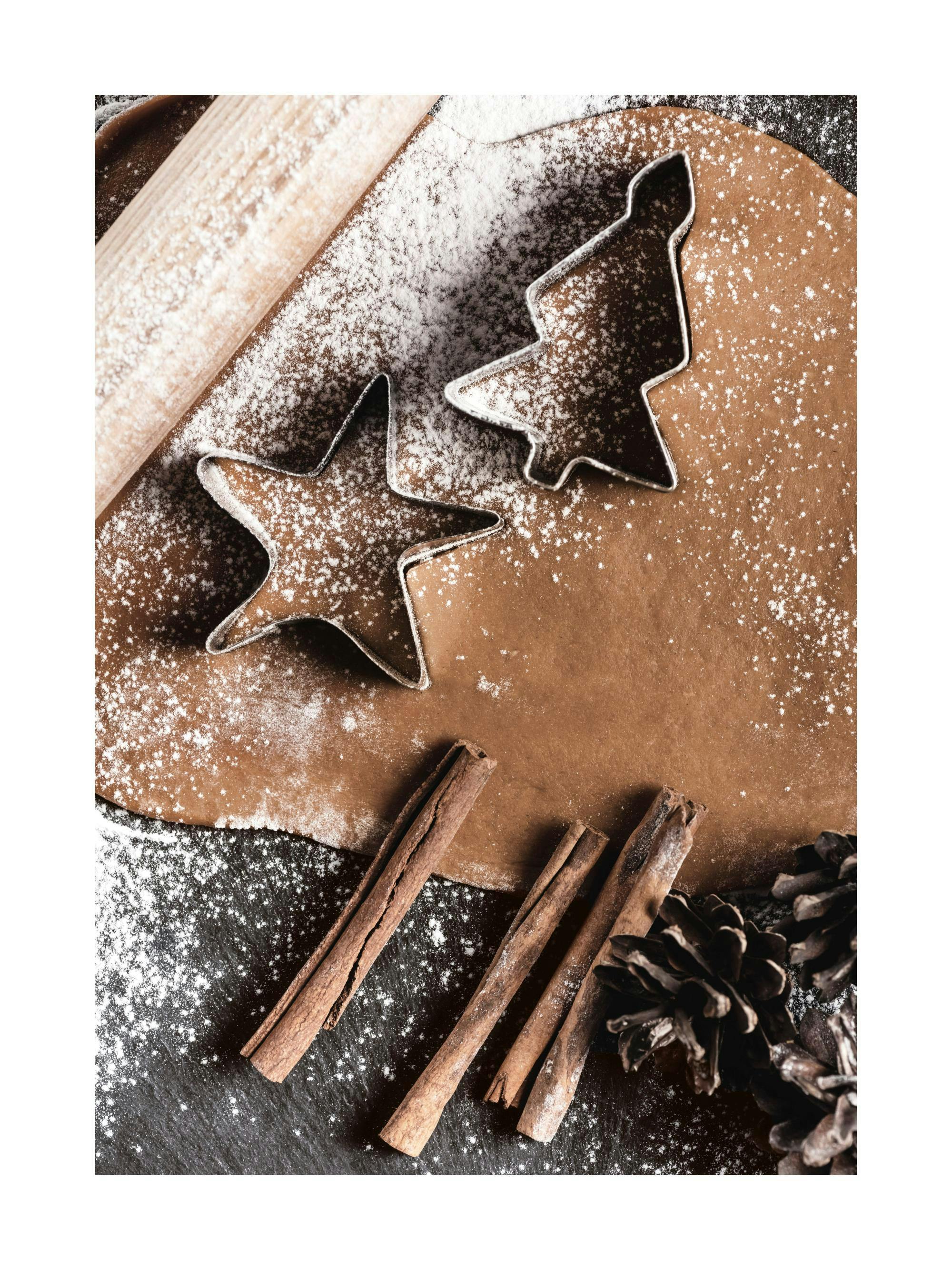 Poster con impasto per biscotti, mattarello, formine, cannella e pigne su sfondo scuro.