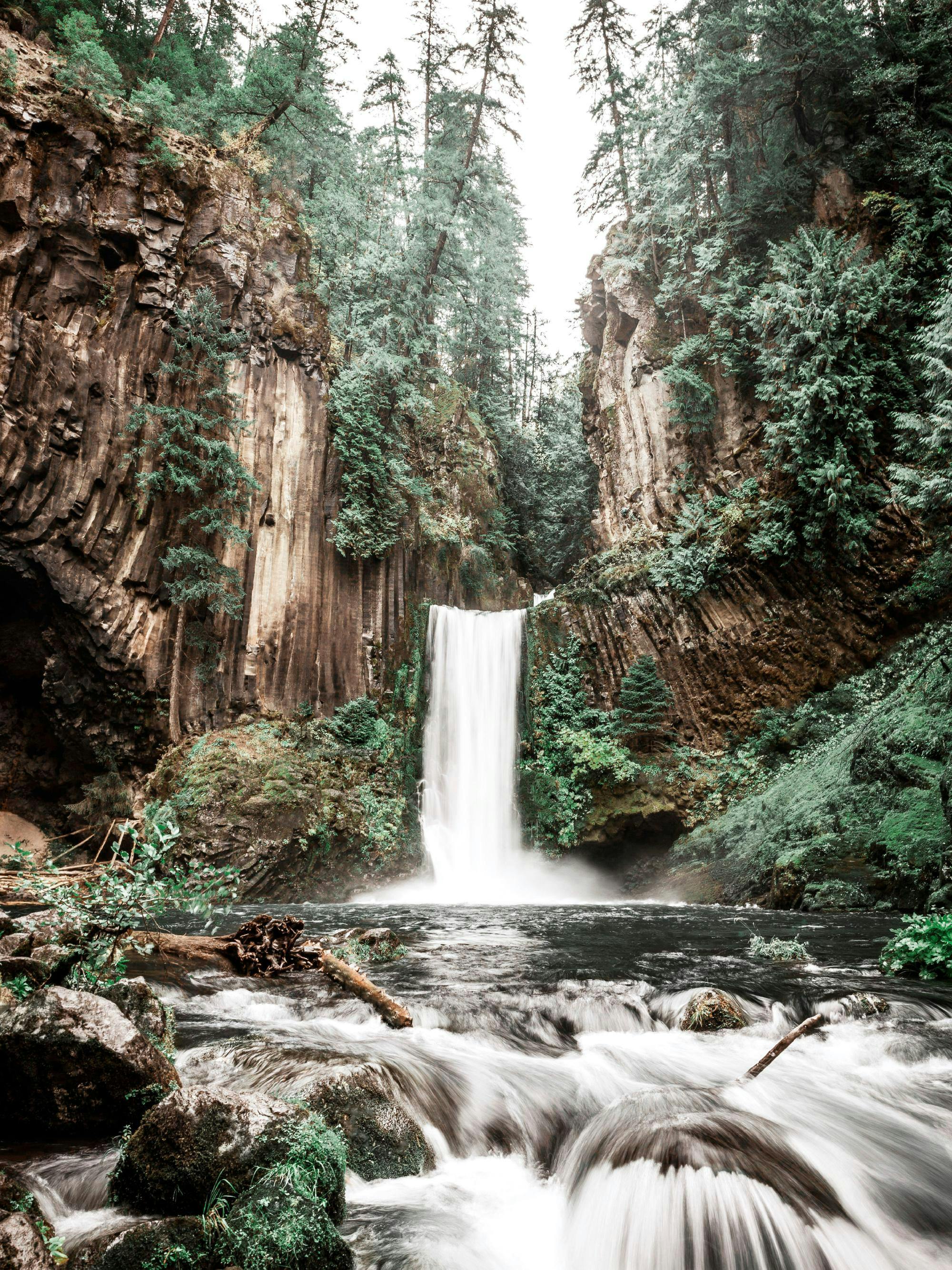 Poster cu o cascadă înaltă, înconjurată de pereți stâncoși și păduri de conifere, cu un râu ce curge în prim plan.