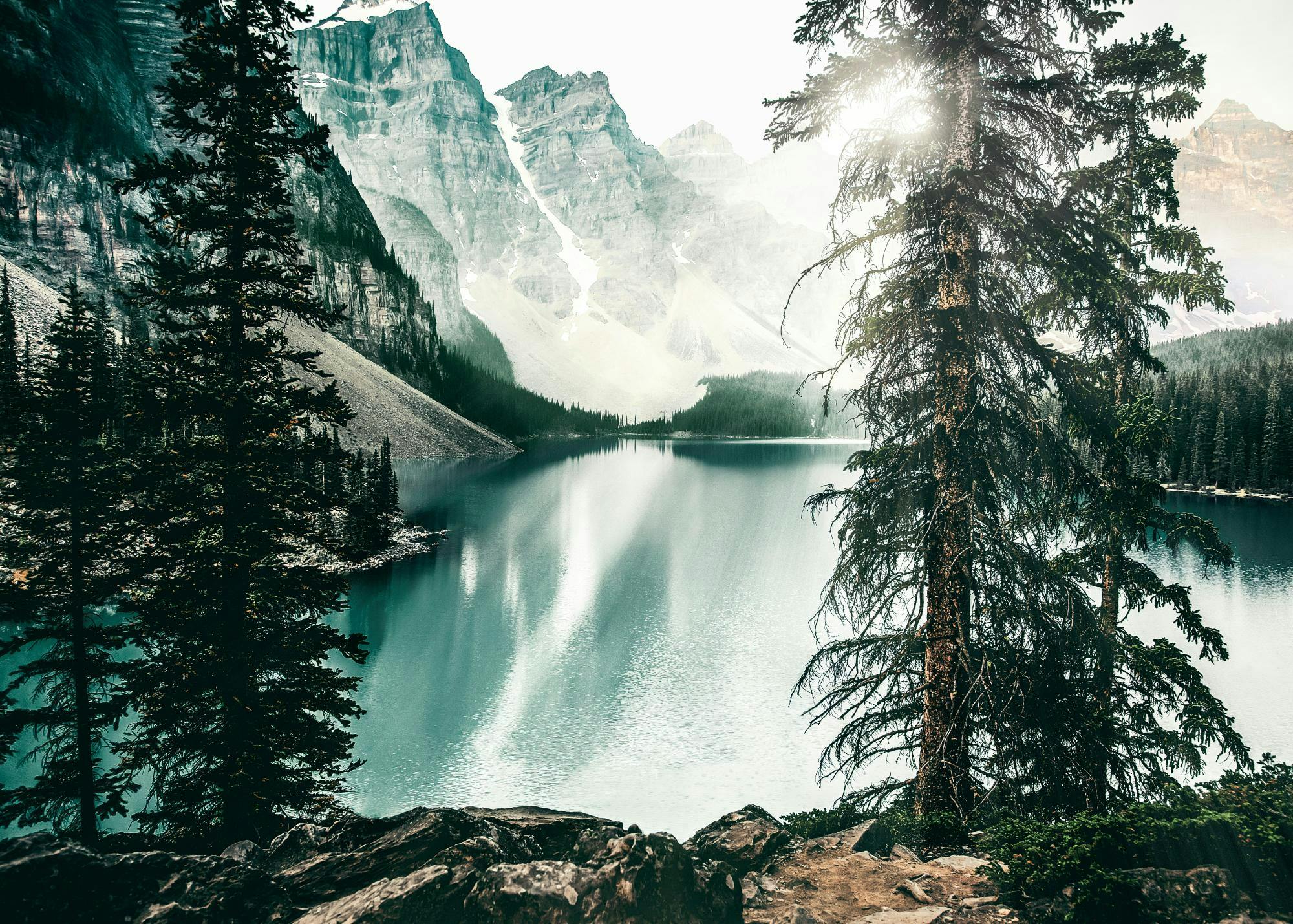 Un poster di un lago alpino con acqua turchese e montagne rocciose, con alberi in primo piano e riflessi solari.