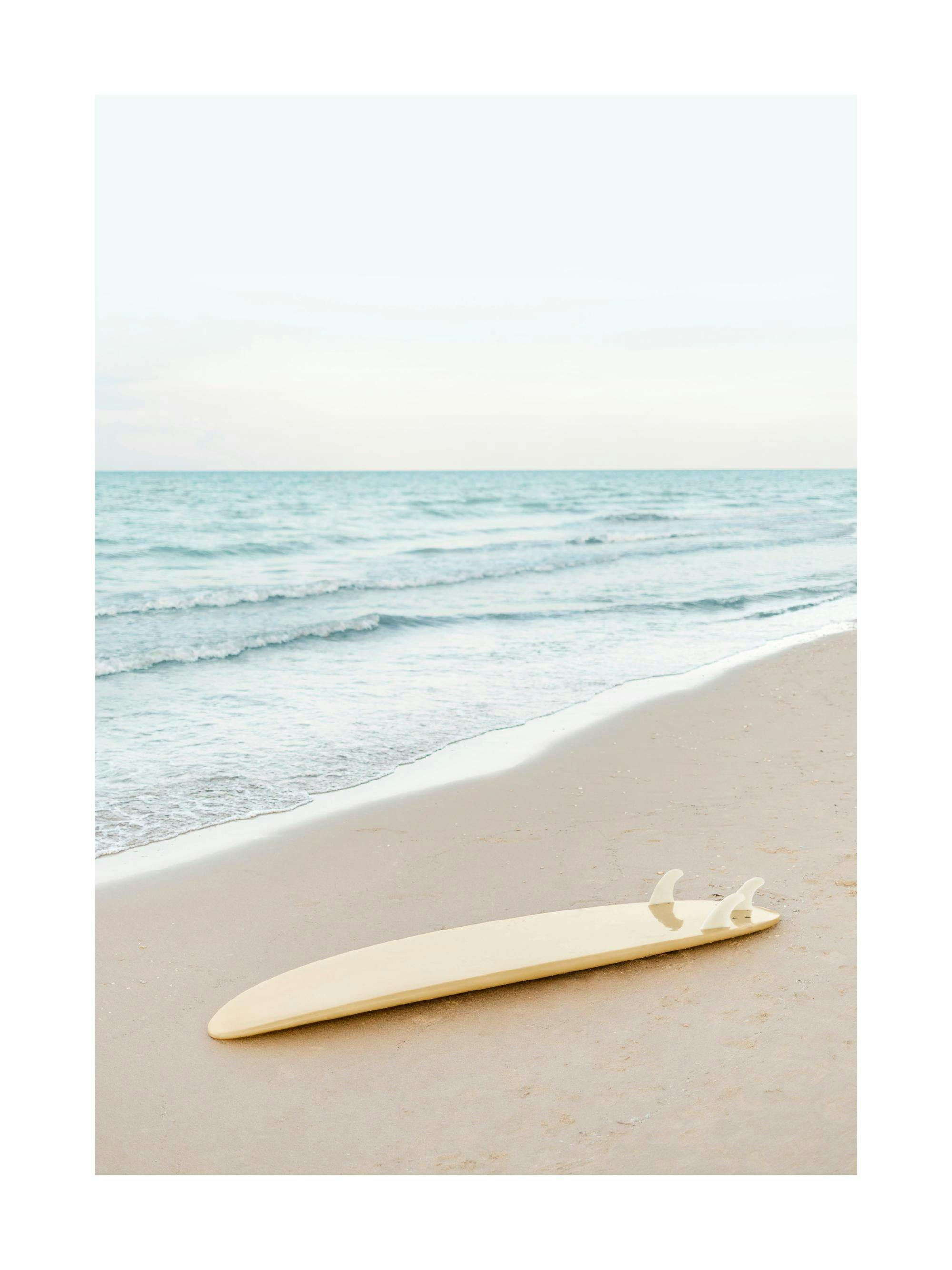 Plakat przedstawiający beżową deskę surfingową leżącą na piaszczystej plaży obok falującego morza.