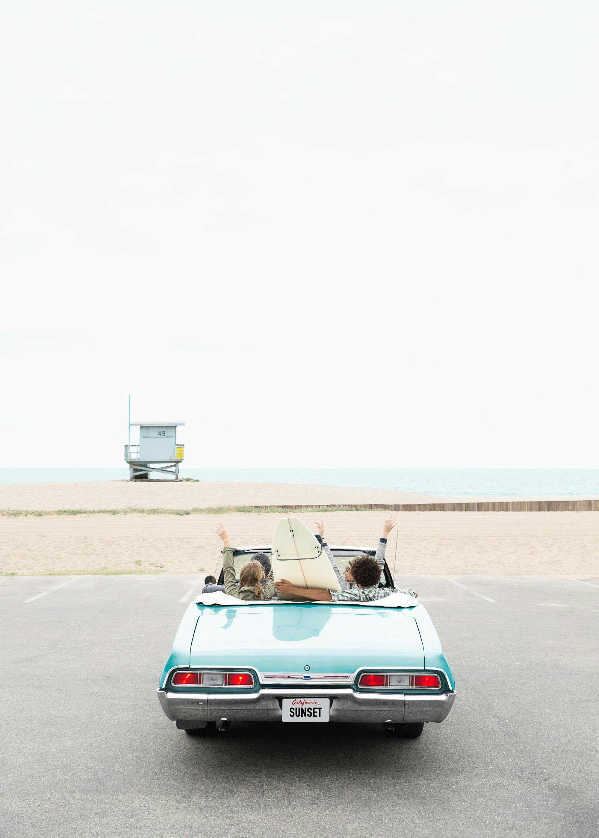 A poster featuring a vintage teal convertible with two people and a surfboard, parked facing a beach with a lifeguard stand.