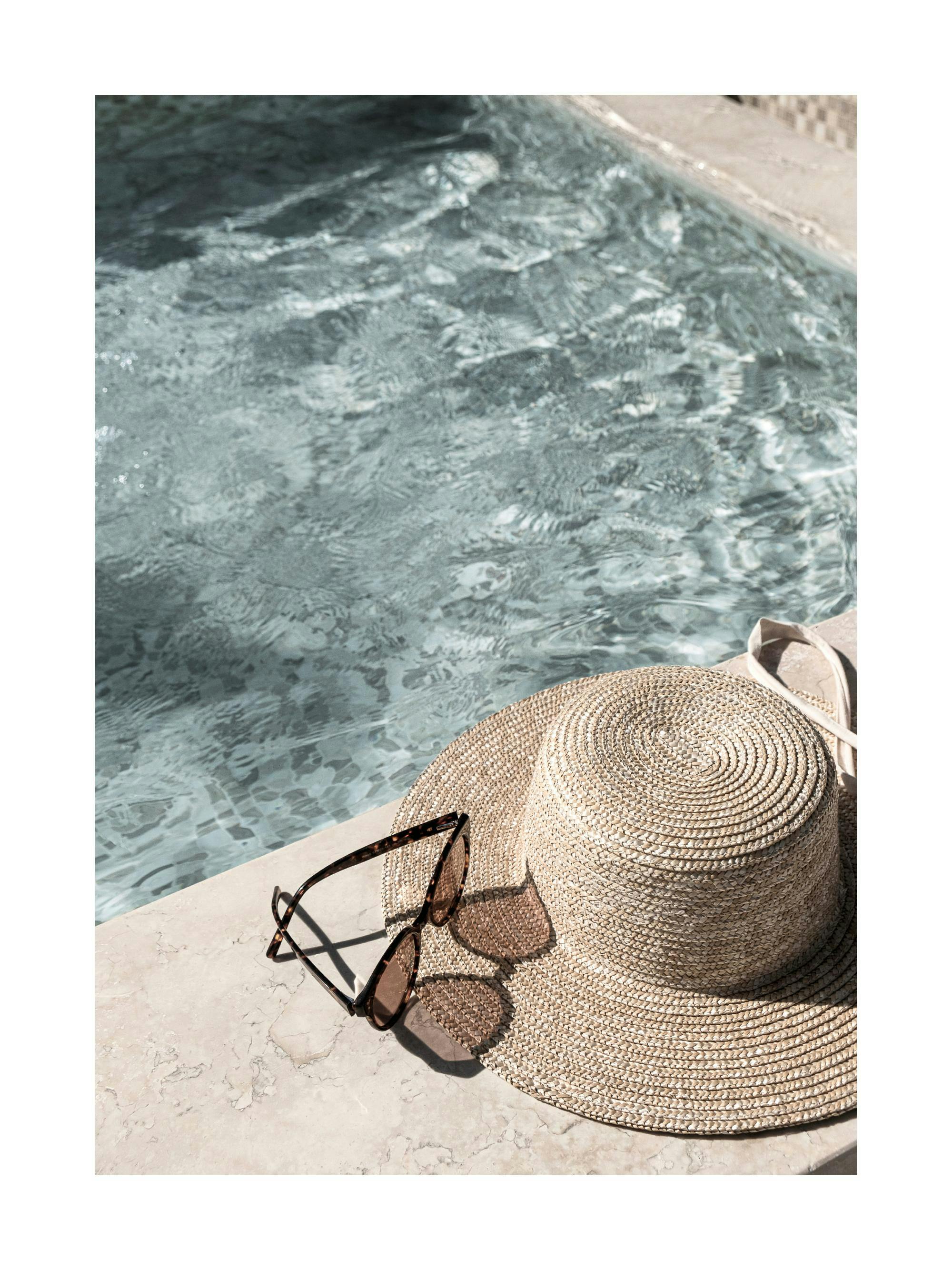 Plakat przedstawia kapelusz słomkowy i okulary przeciwsłoneczne na skraju basenu o turkusowej, falującej wodzie.