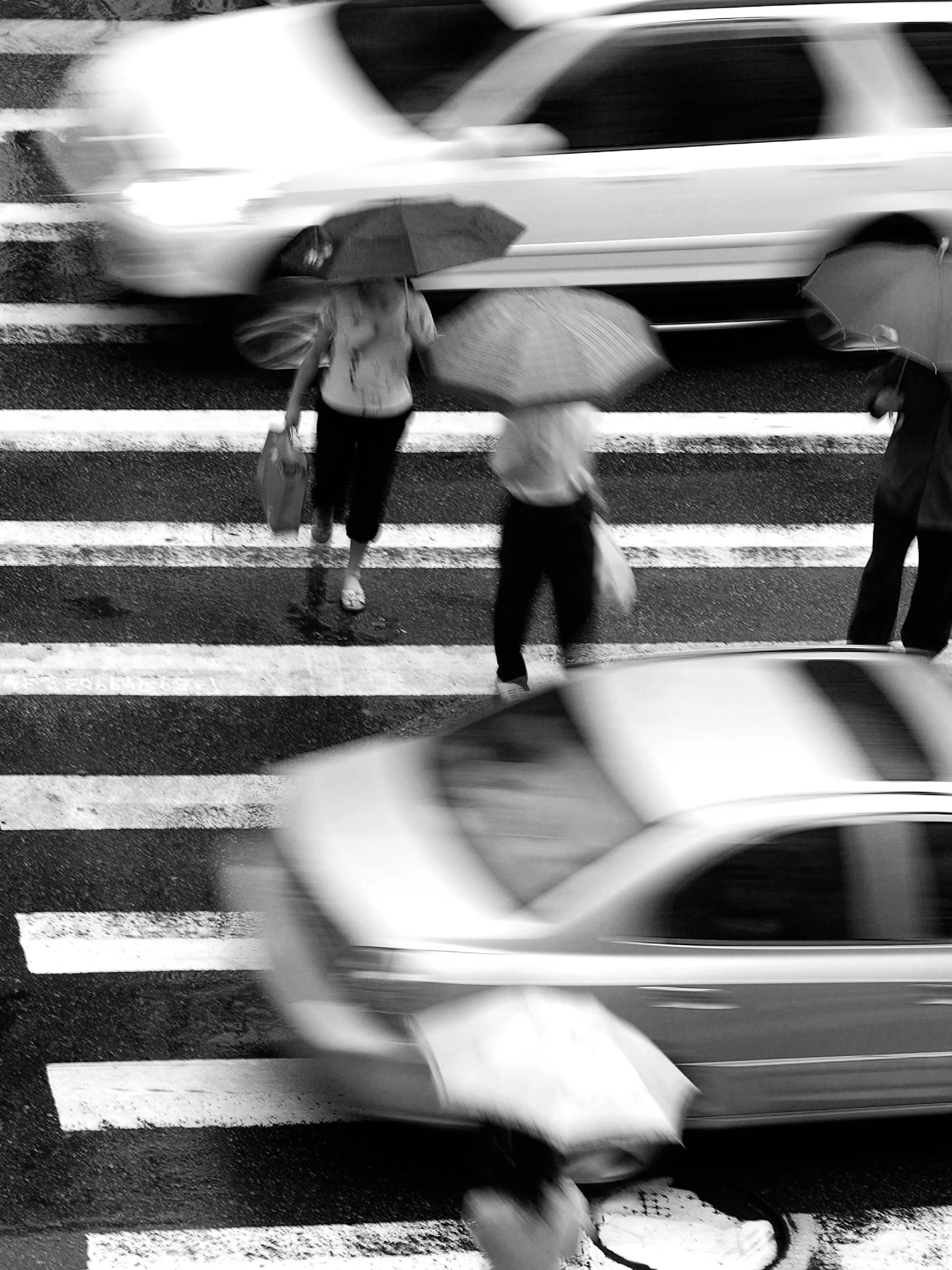 A poster featuring a black and white blurred street scene with people holding umbrellas crossing a crosswalk as cars drive by.