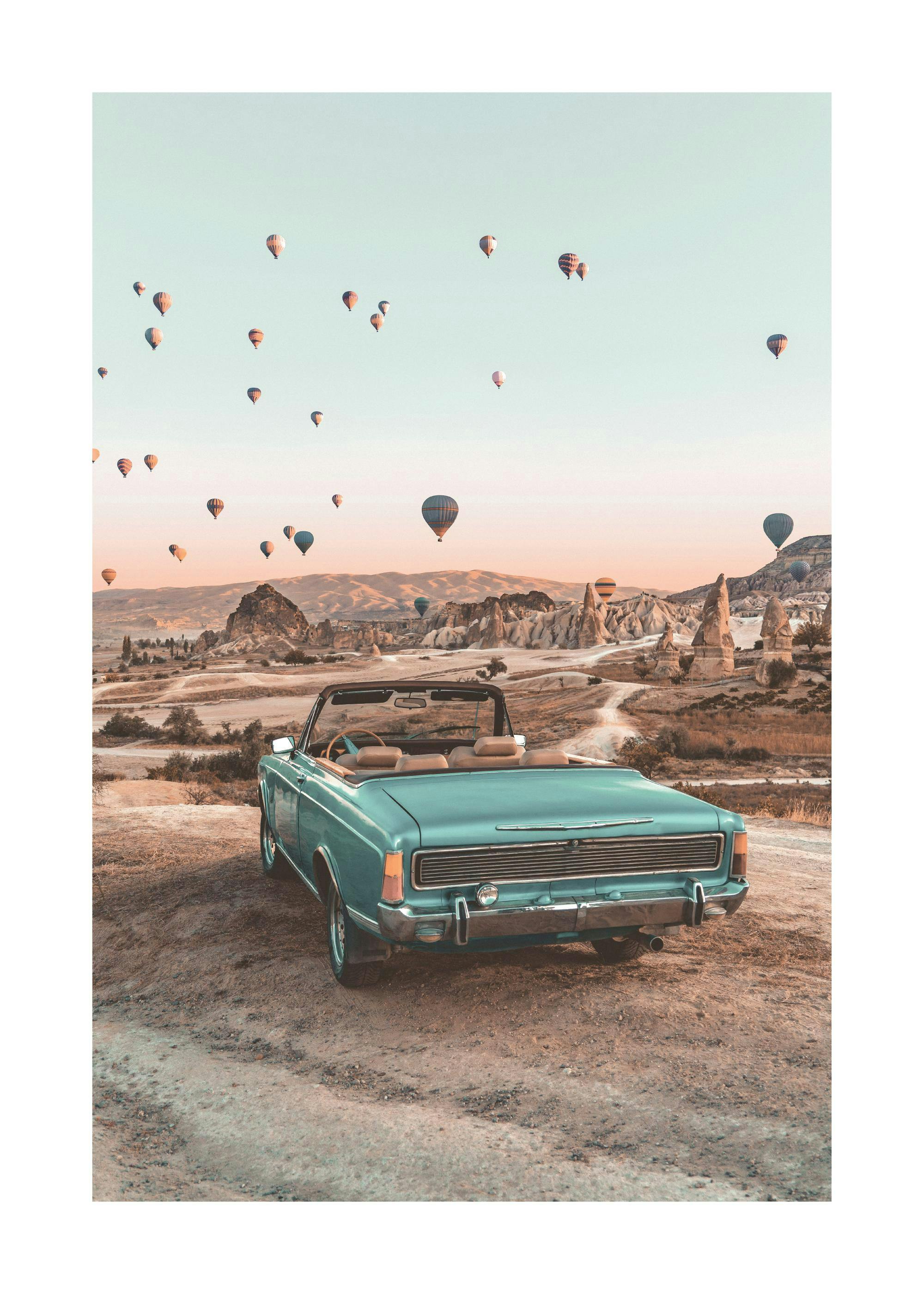 A poster of a vintage teal convertible car parked on a dirt road, overlooking a valley filled with hot air balloons at sunset.