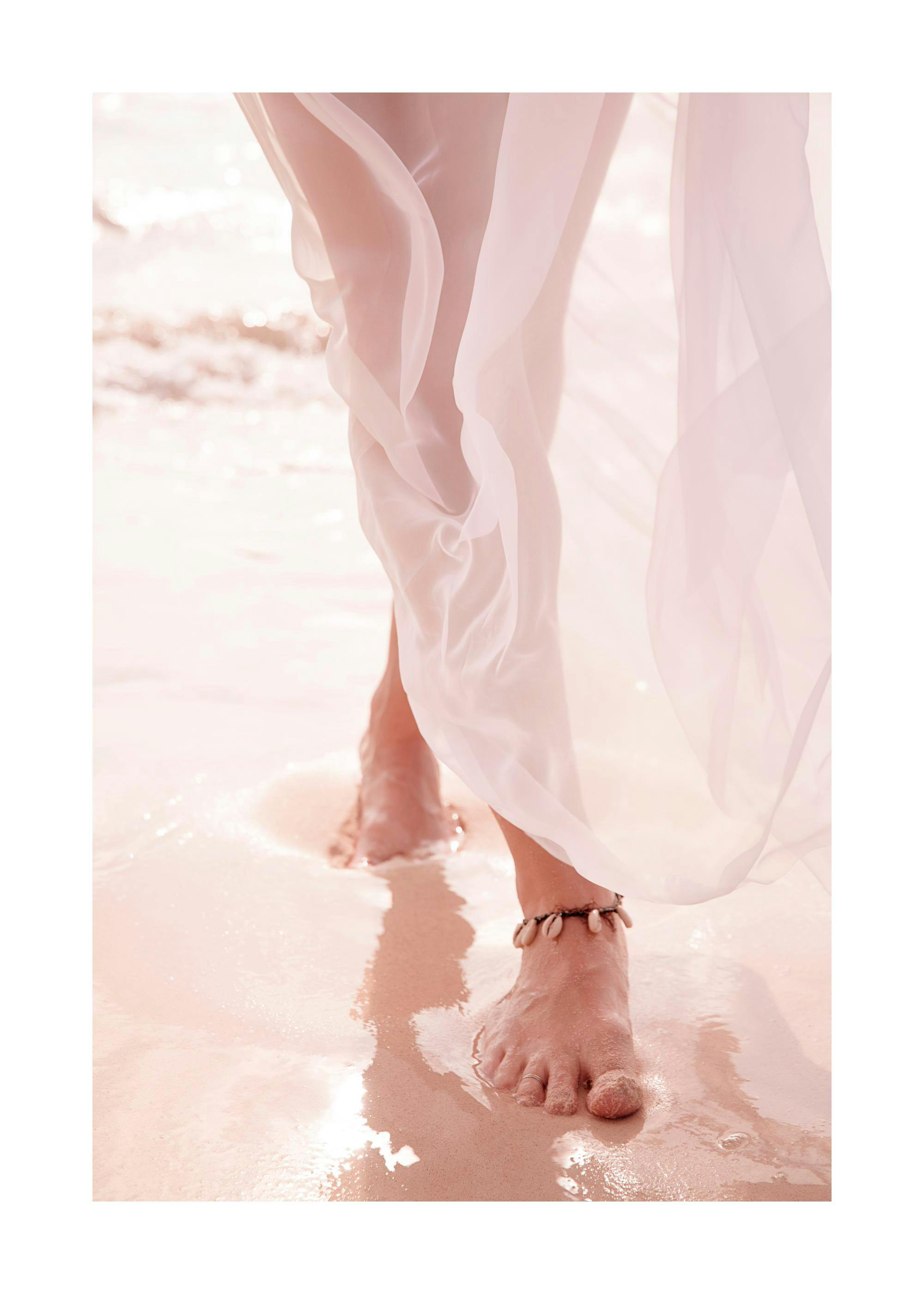 A poster featuring a womans bare feet walking in shallow water on a sandy beach, with a sheer white fabric flowing around her le