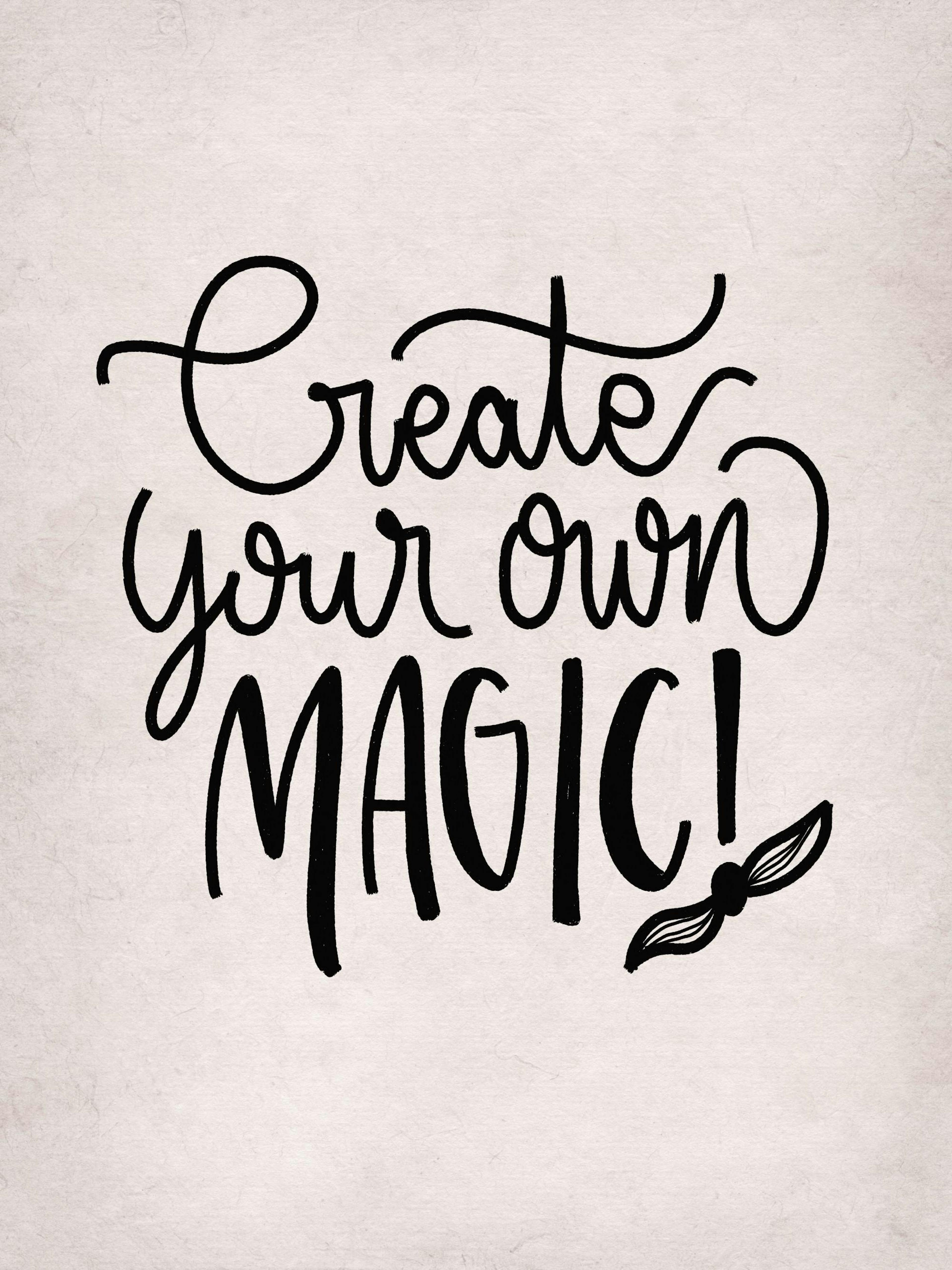 A poster with the black handwritten text Create your own MAGIC! in a light wood frame, displayed on a wooden desk with a lamp an
