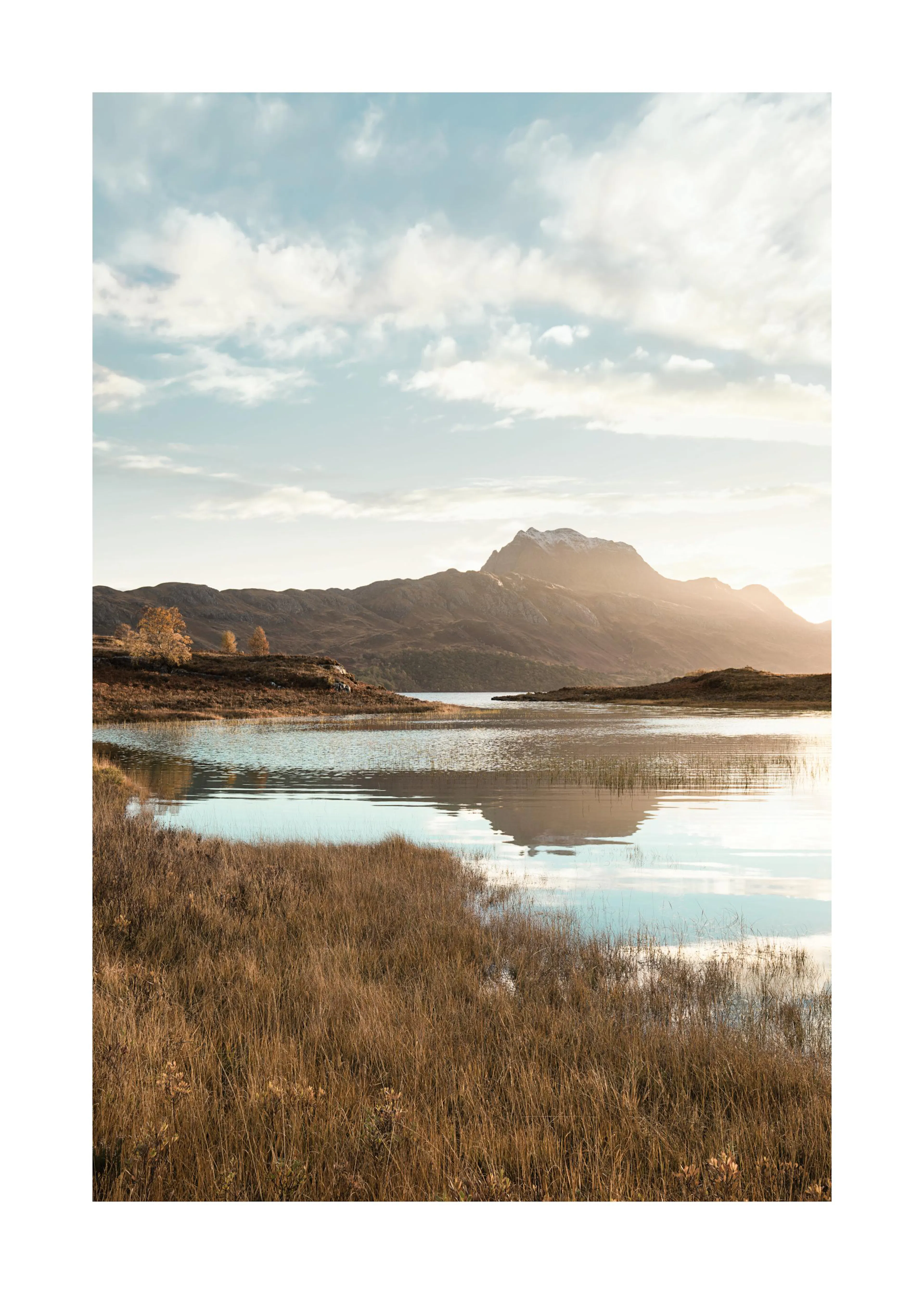 Poster con un lago tranquillo e montagne sullo sfondo, erba secca in primo piano e cielo nuvoloso al tramonto.