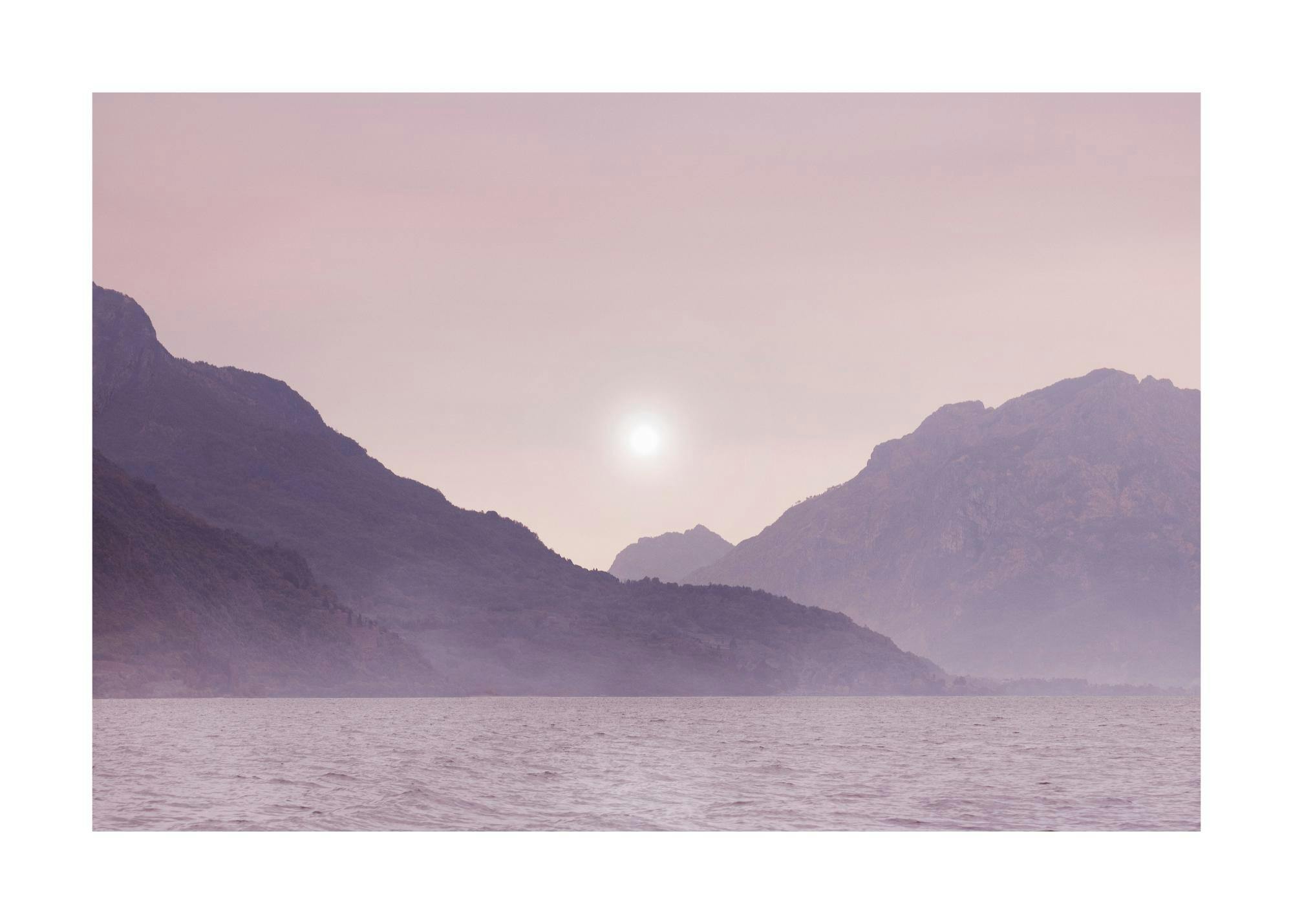 Póster de un lago tranquilo con montañas oscuras y un sol brillante en un cielo brumoso de color lavanda.