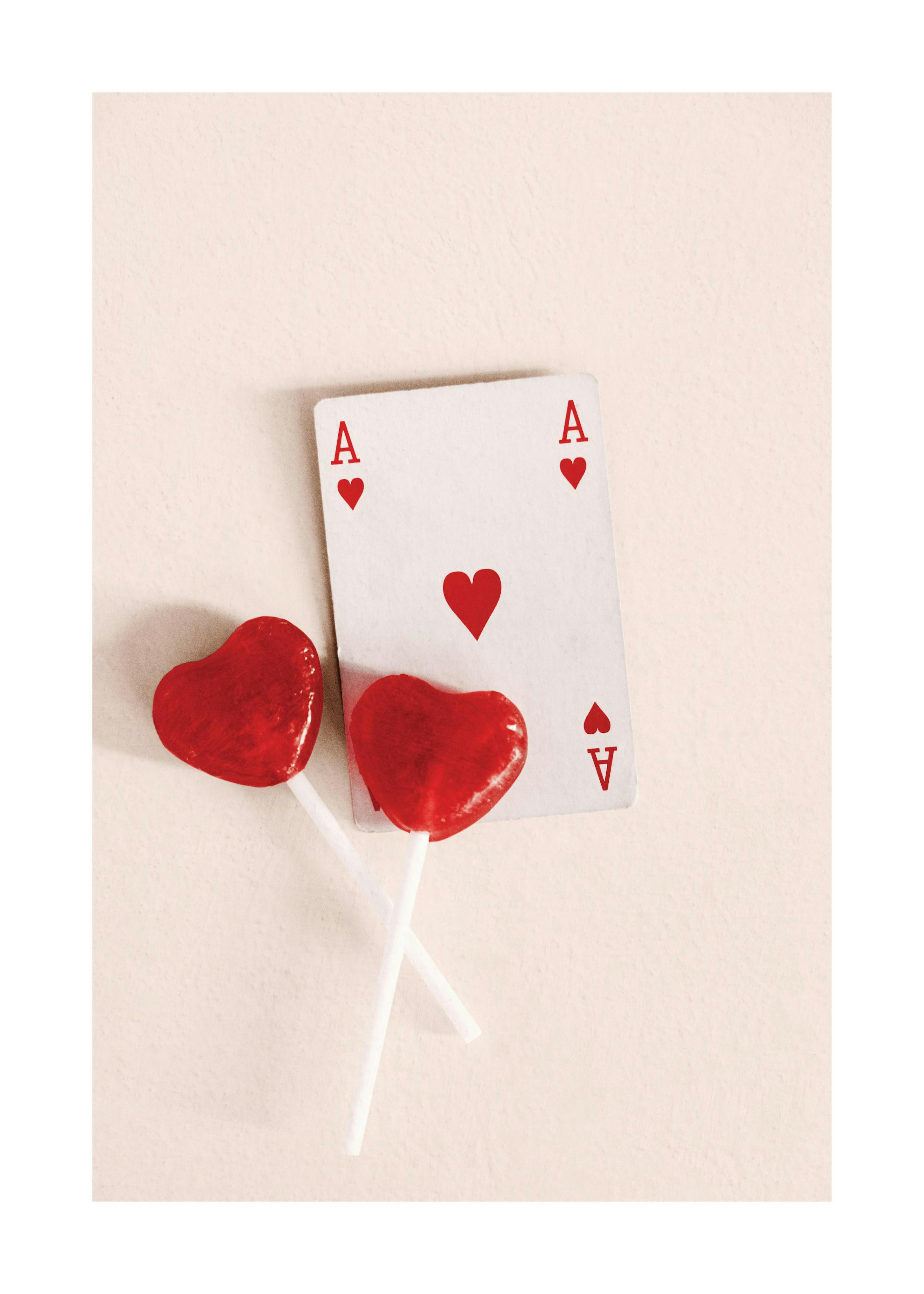 A poster featuring a white Ace of Hearts playing card alongside two red heart-shaped lollipops on a light background.