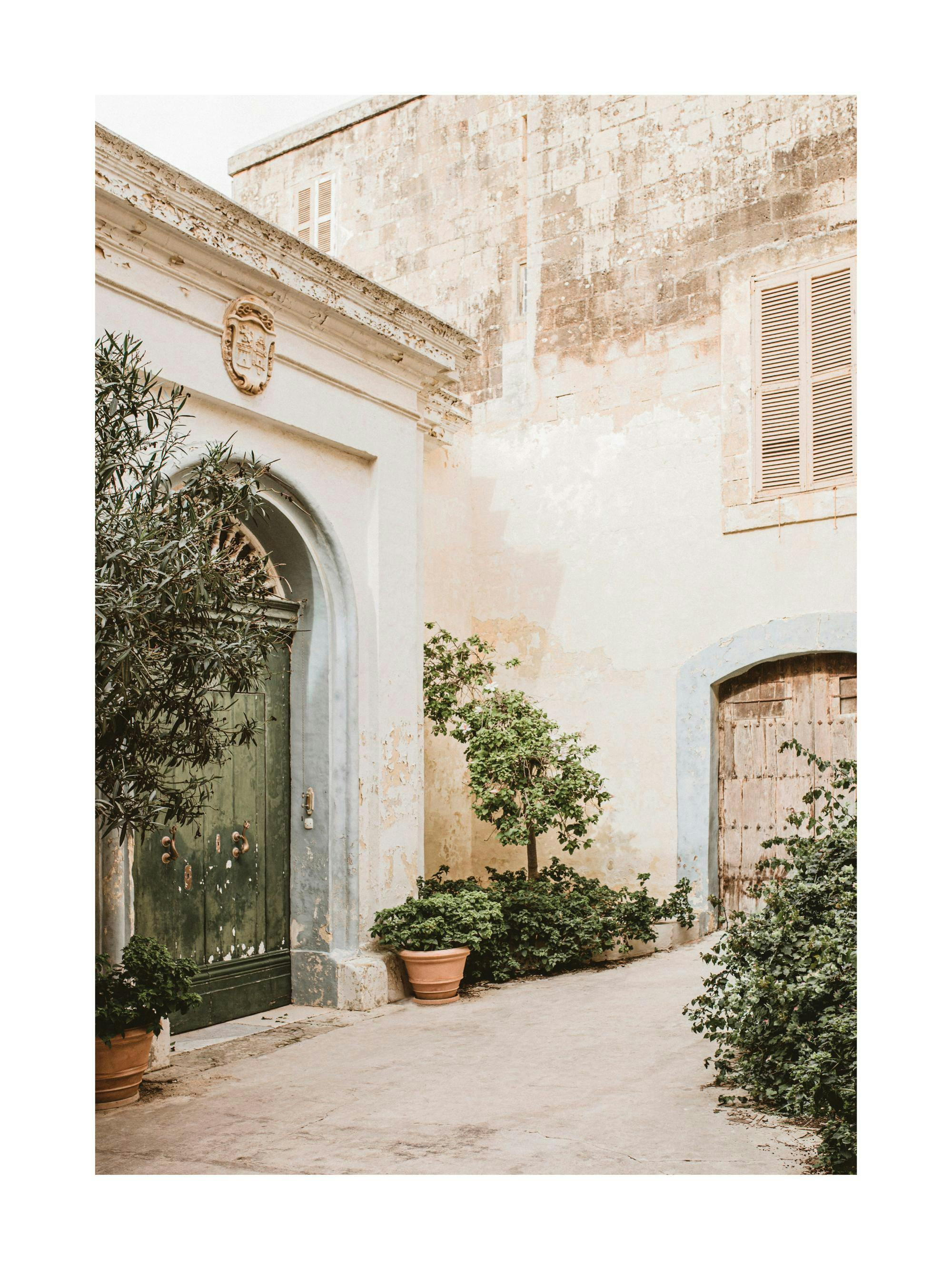 A poster featuring a sun-drenched, rustic European street with aged stone walls, old doors and lush green plants.