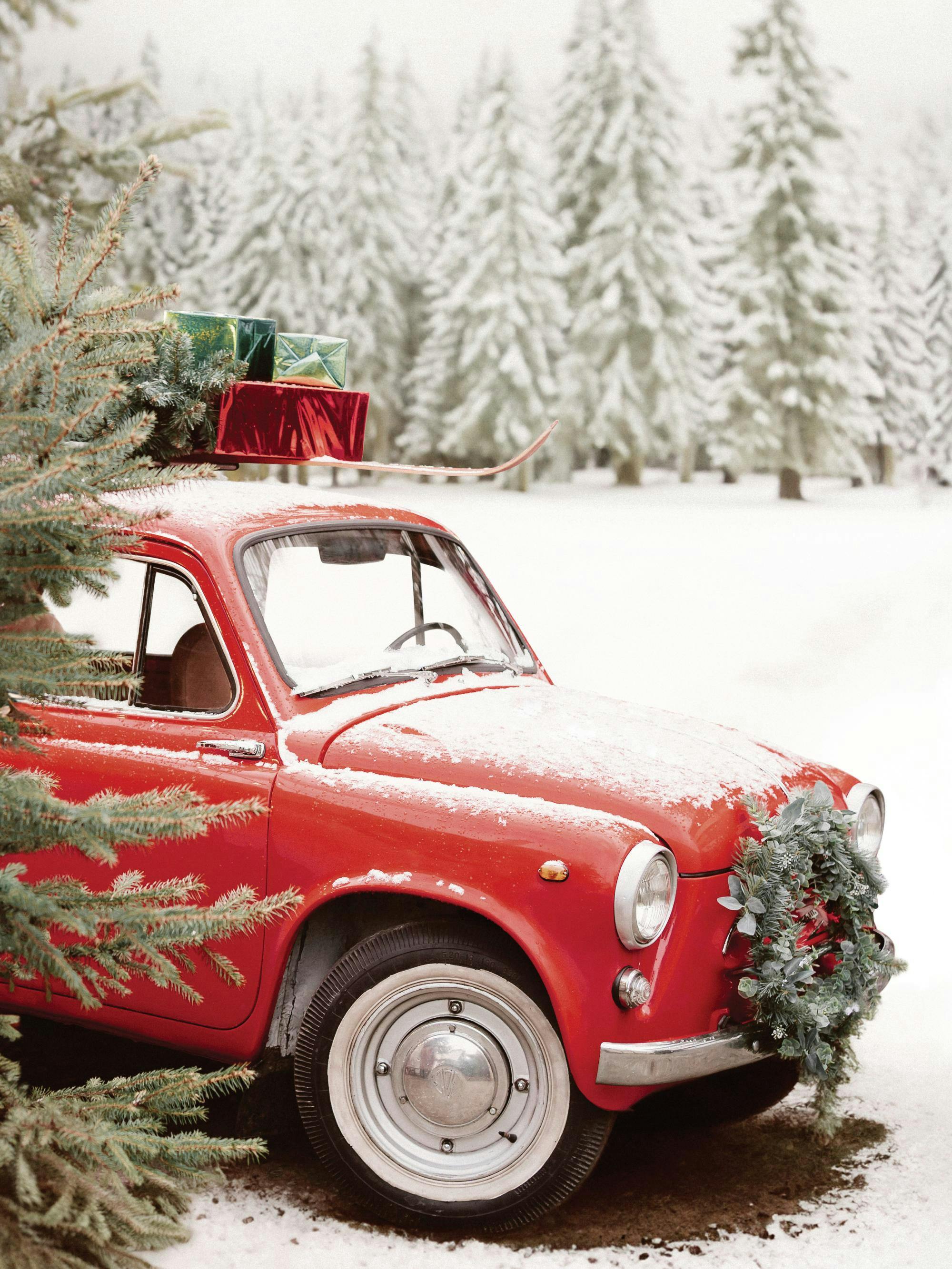 Jouluaiheinen juliste: Punainen retroauto lumisessa maastossa lähellä kuusia, auton katolla lahjoja.