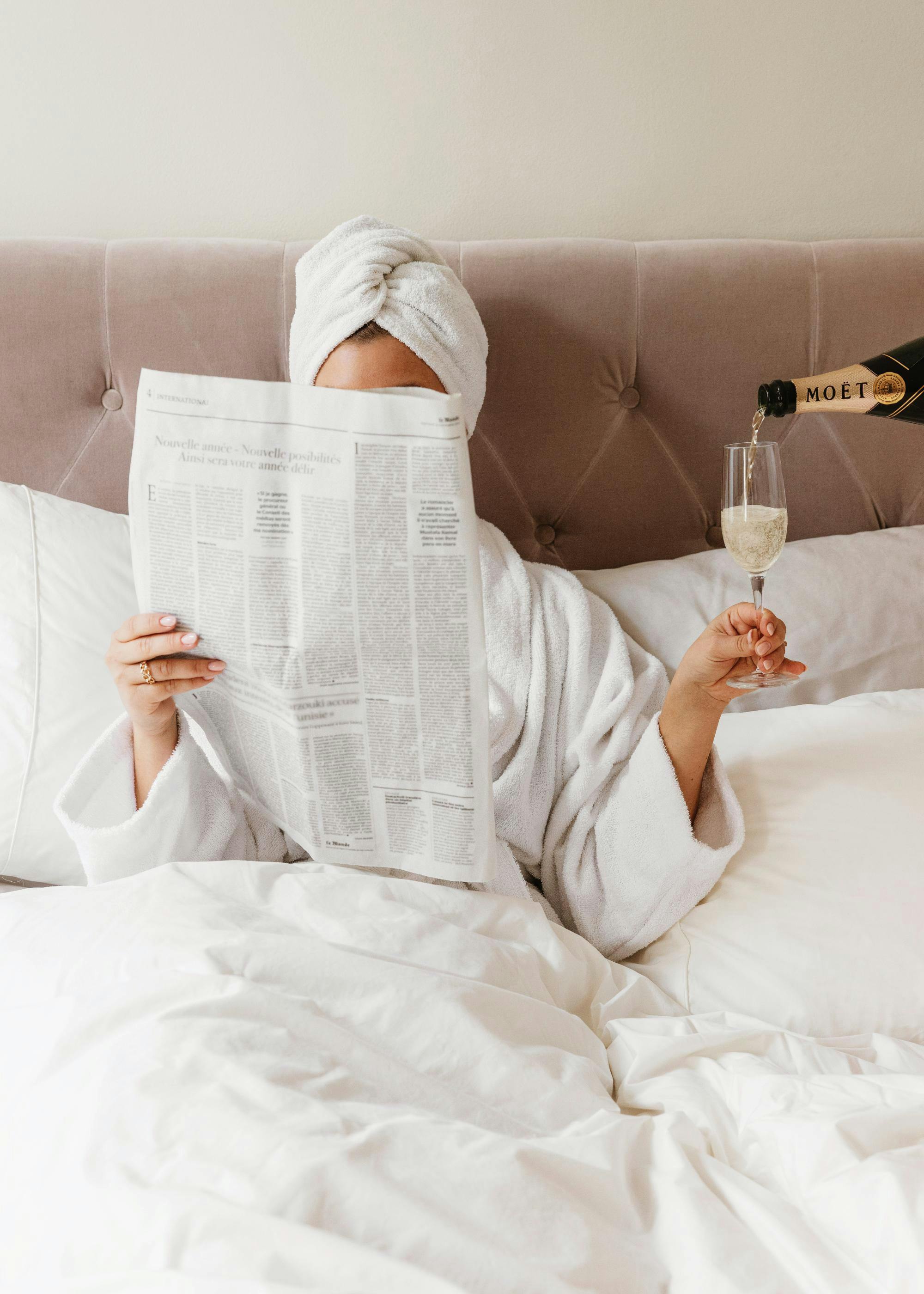 A poster featuring a person in a bathrobe and towel turban, reading a newspaper in bed while champagne is poured into a glass.