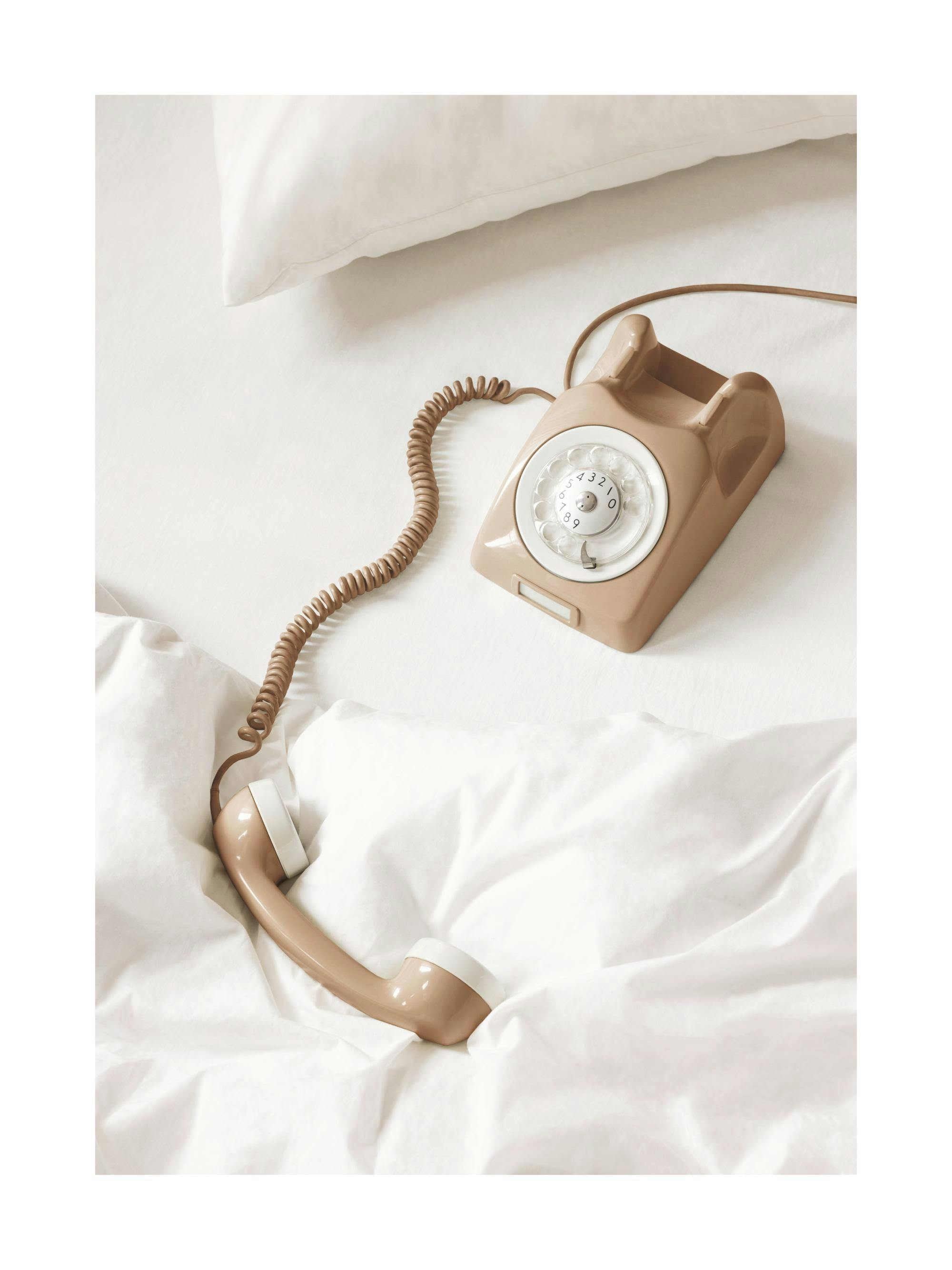 Póster de un teléfono retro beige con dial giratorio en una cama blanca, con el auricular descolgado.