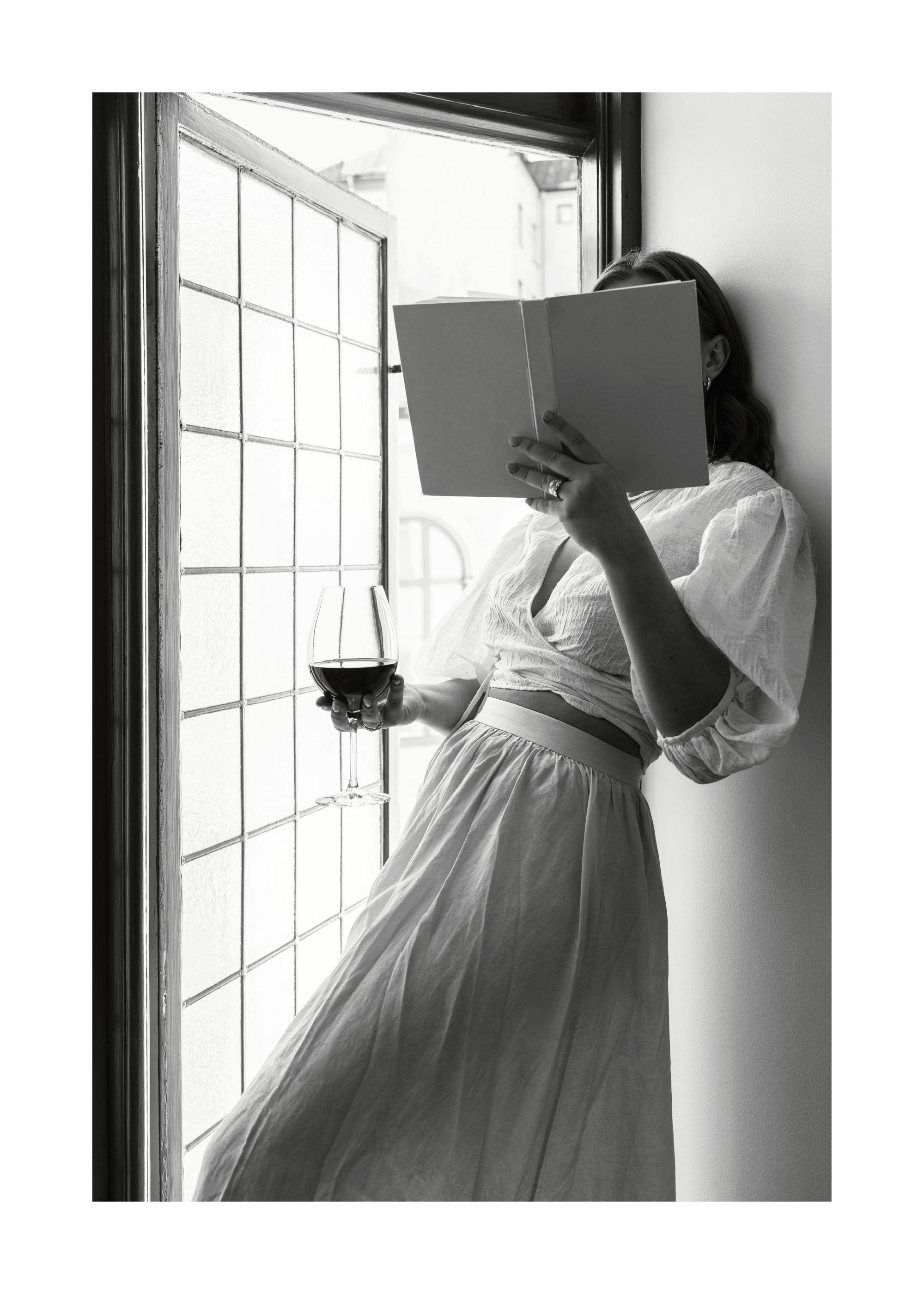 Poster noir et blanc dune personne lisant un livre, un verre de vin à la main, près dune fenêtre à petits carreaux.