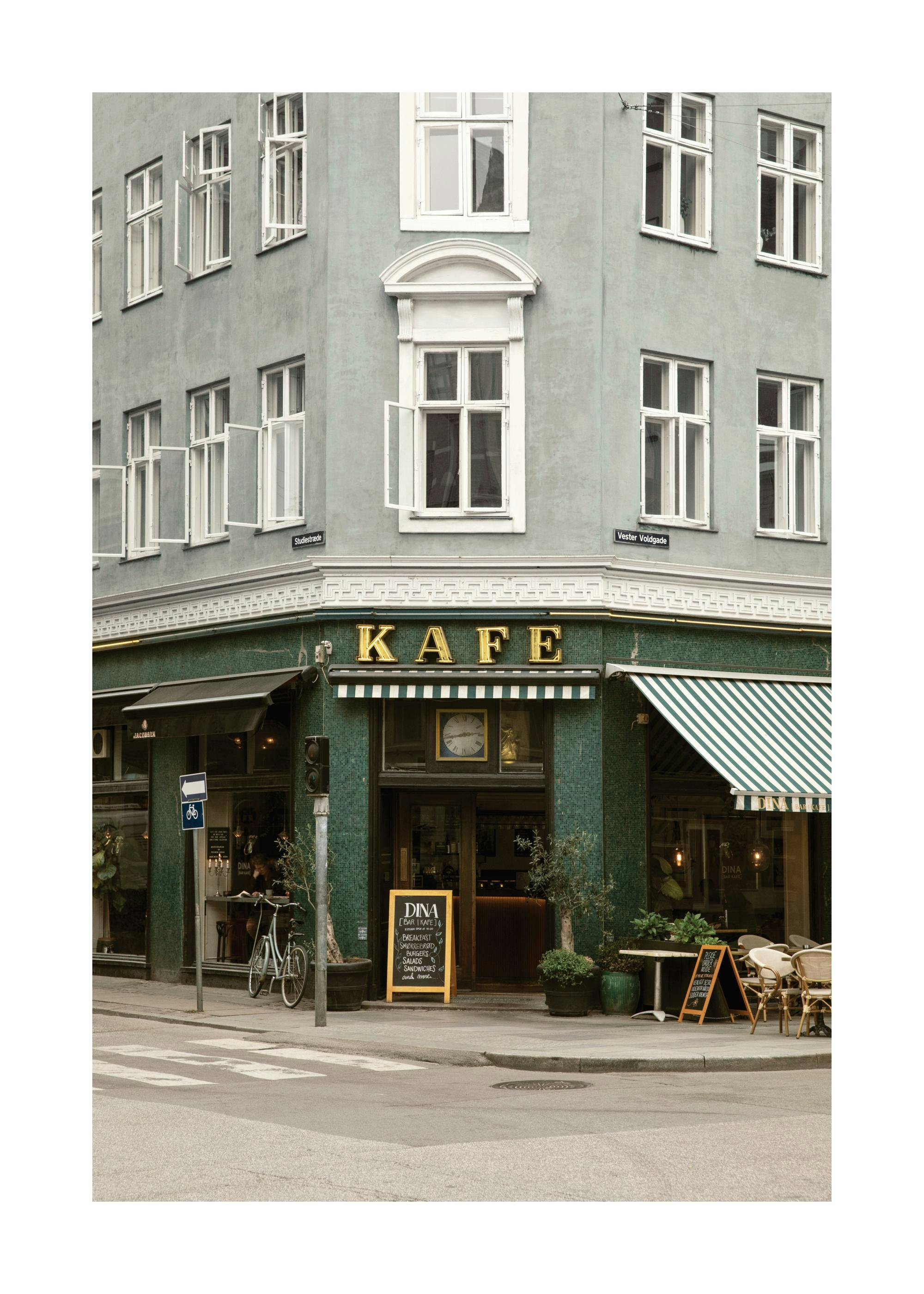 Póster de un café de esquina con toldos de rayas verdes y blancas, sillas de mimbre y el letrero KAFE en dorado.