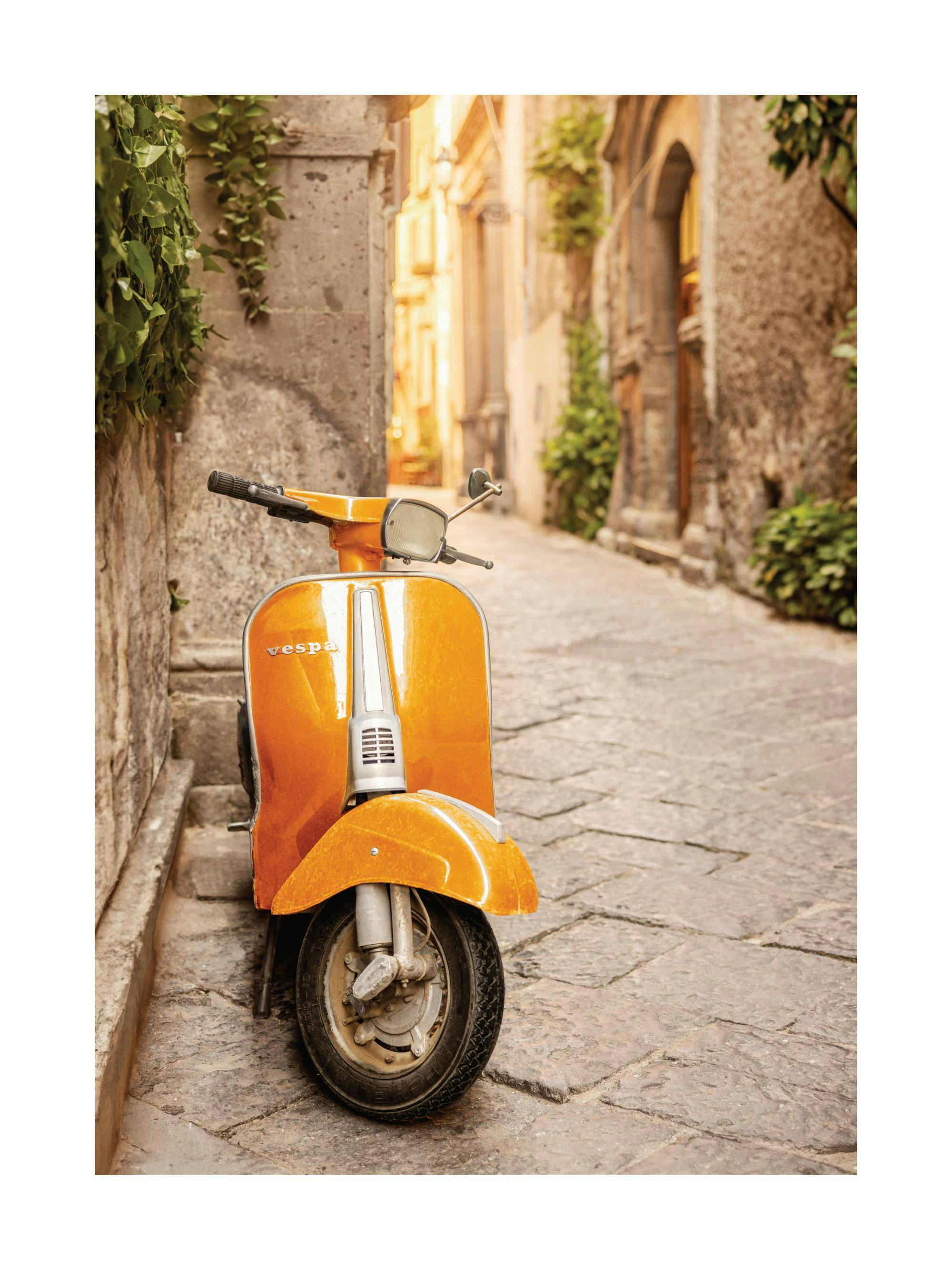 Plakát s oranžovou Vespou zaparkovanou na dlážděné ulici mezi starými kamennými domy, s popínavými rostlinami.