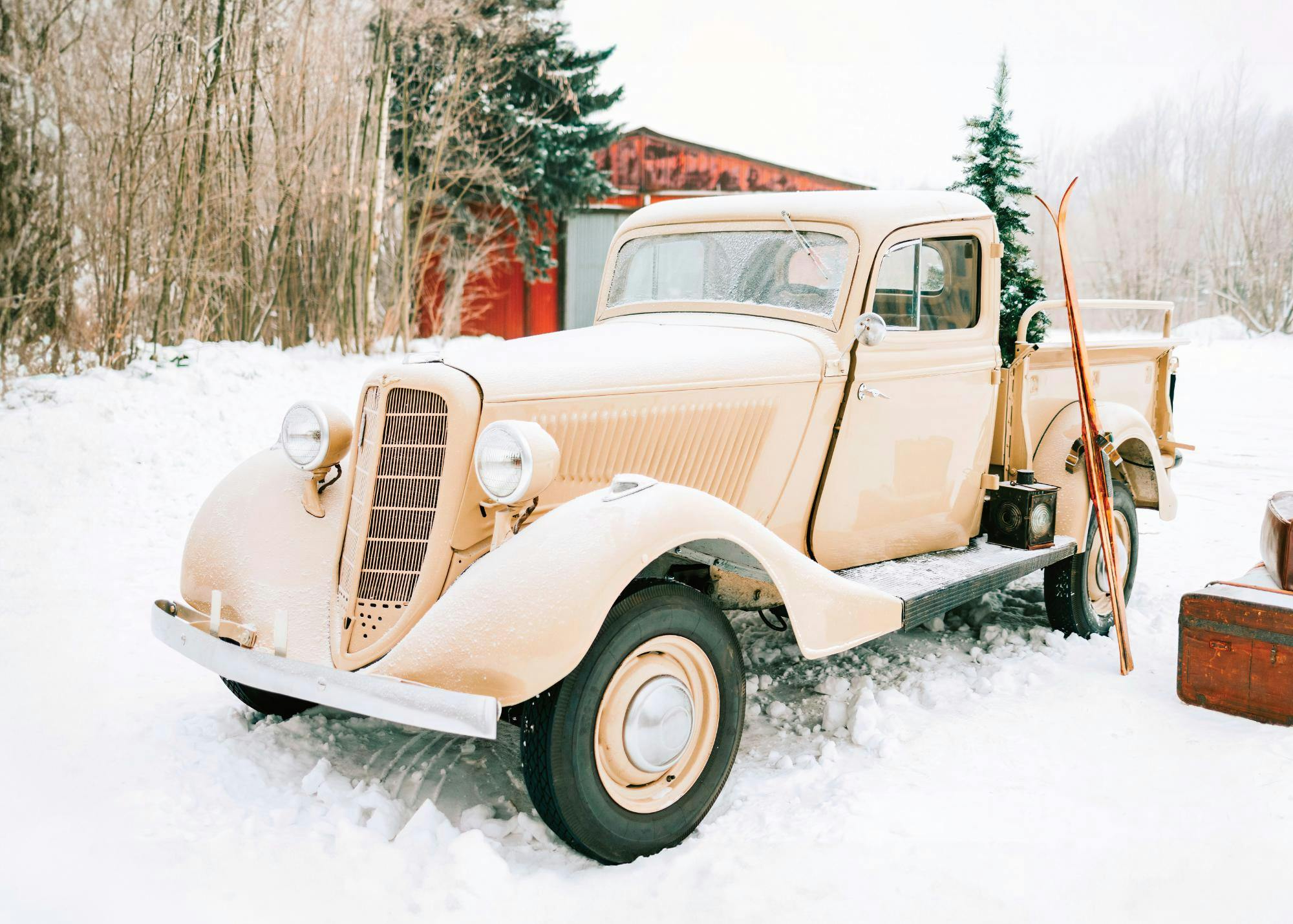 A vintage beige truck with wooden skis and old suitcases in a snowy landscape, featuring a red barn in the background, on a wint