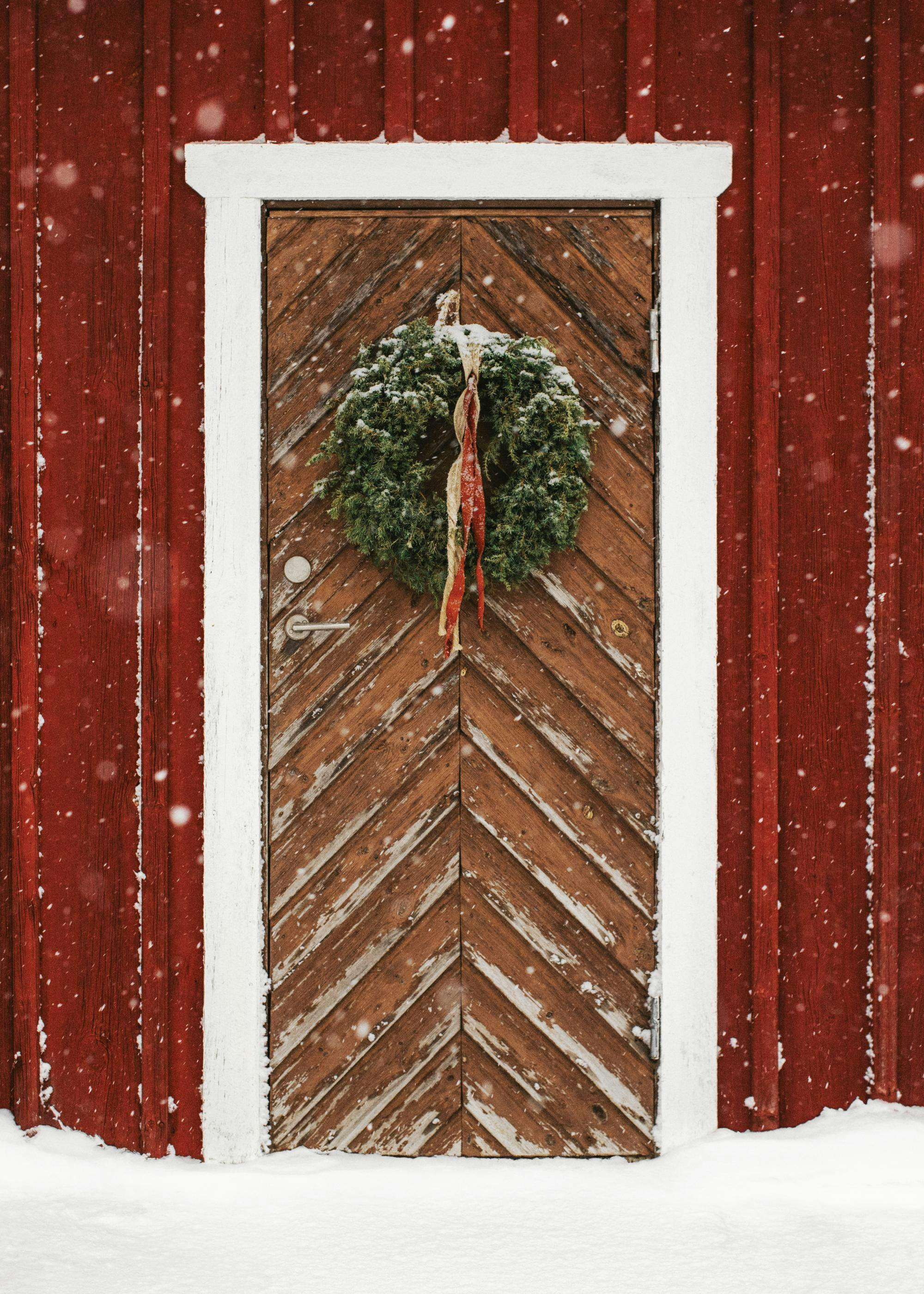 A poster featuring a snowy red house with a brown wooden door and a green wreath with red ribbons.
