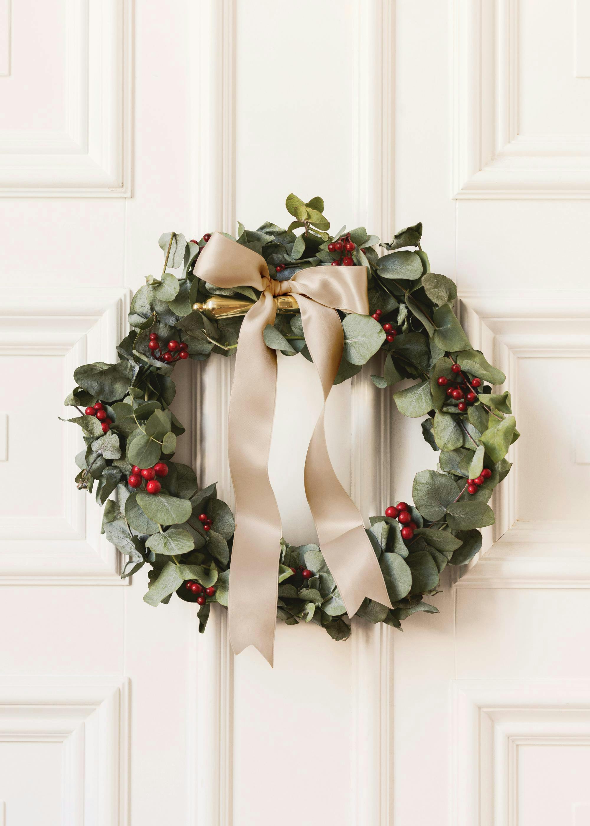A poster of a Christmas wreath with green eucalyptus leaves, red berries, and a gold ribbon hanging on a white door.