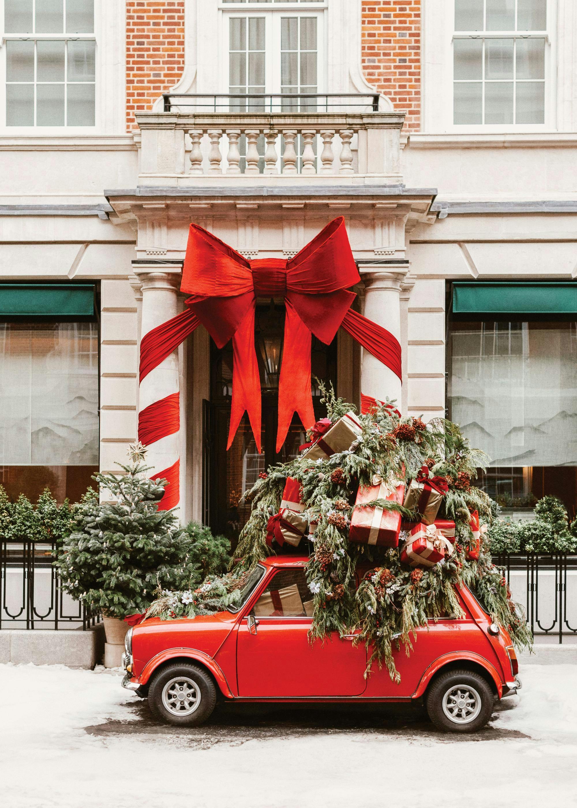 A festive poster featuring a red Mini loaded with Christmas trees and presents, parked outside a building with a large red bow d
