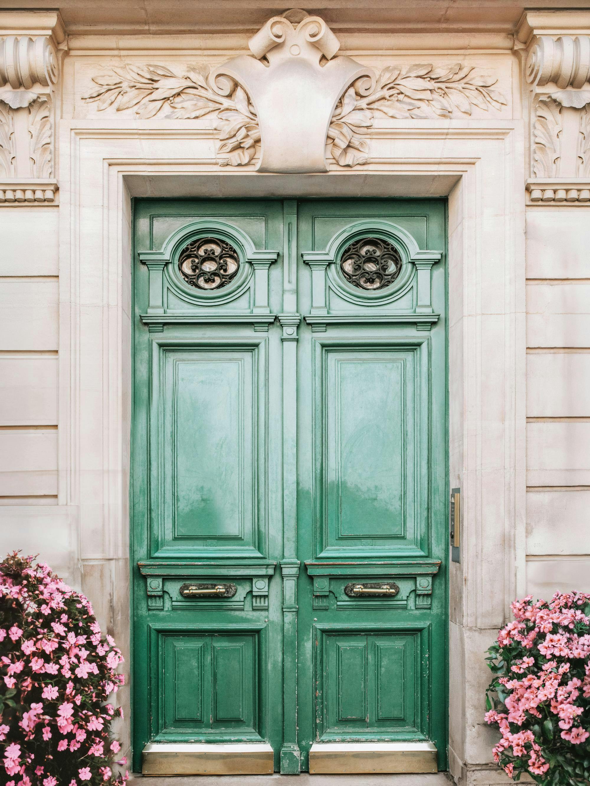 Poster cu o ușă verde veche, cu detalii sculptate, flori roz și un perete din piatră.