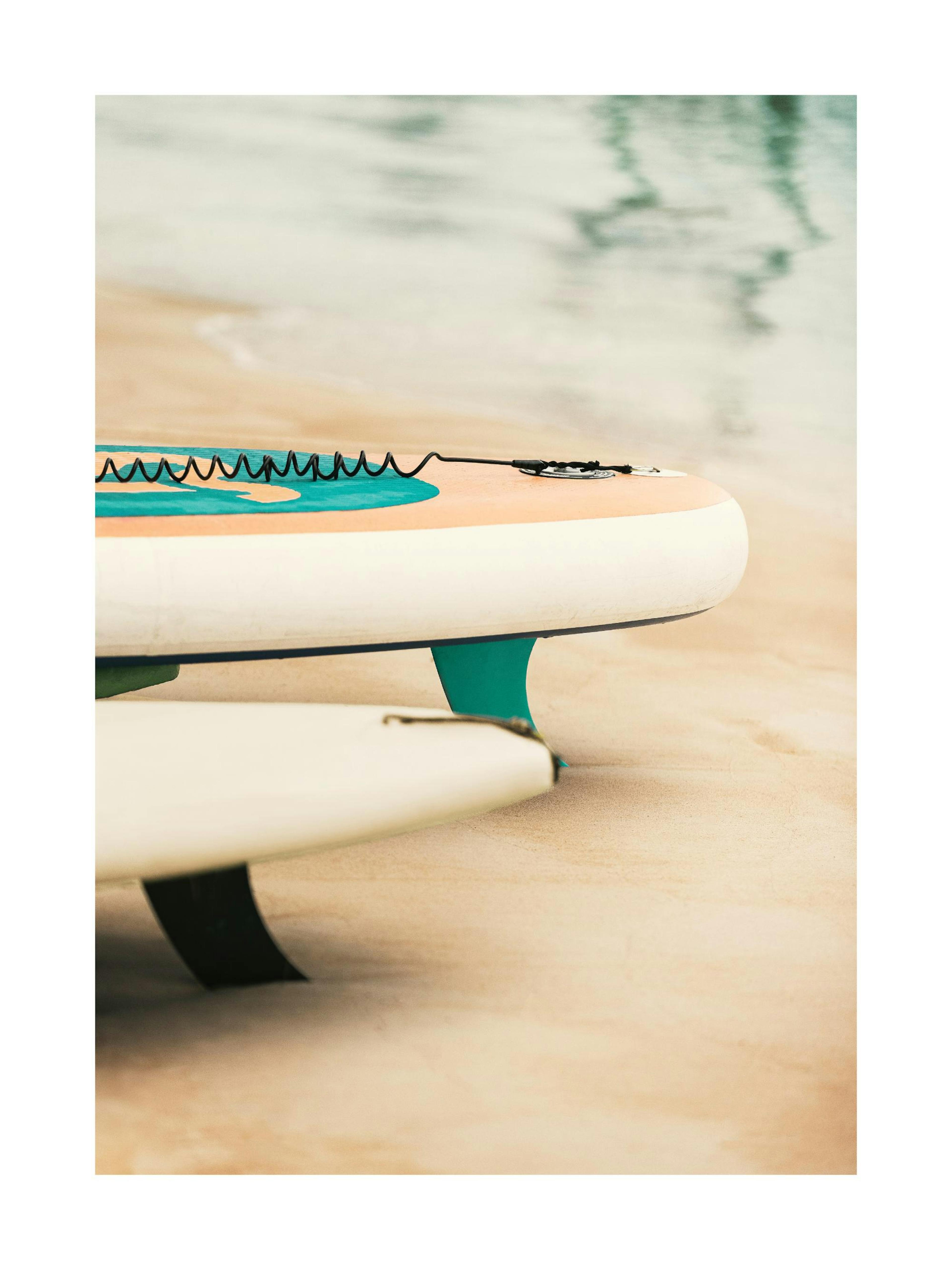 Poster con tavole da surf e paddleboard sulla sabbia chiara, vicino allacqua, esposto su una mensola con oggetti decorativi.