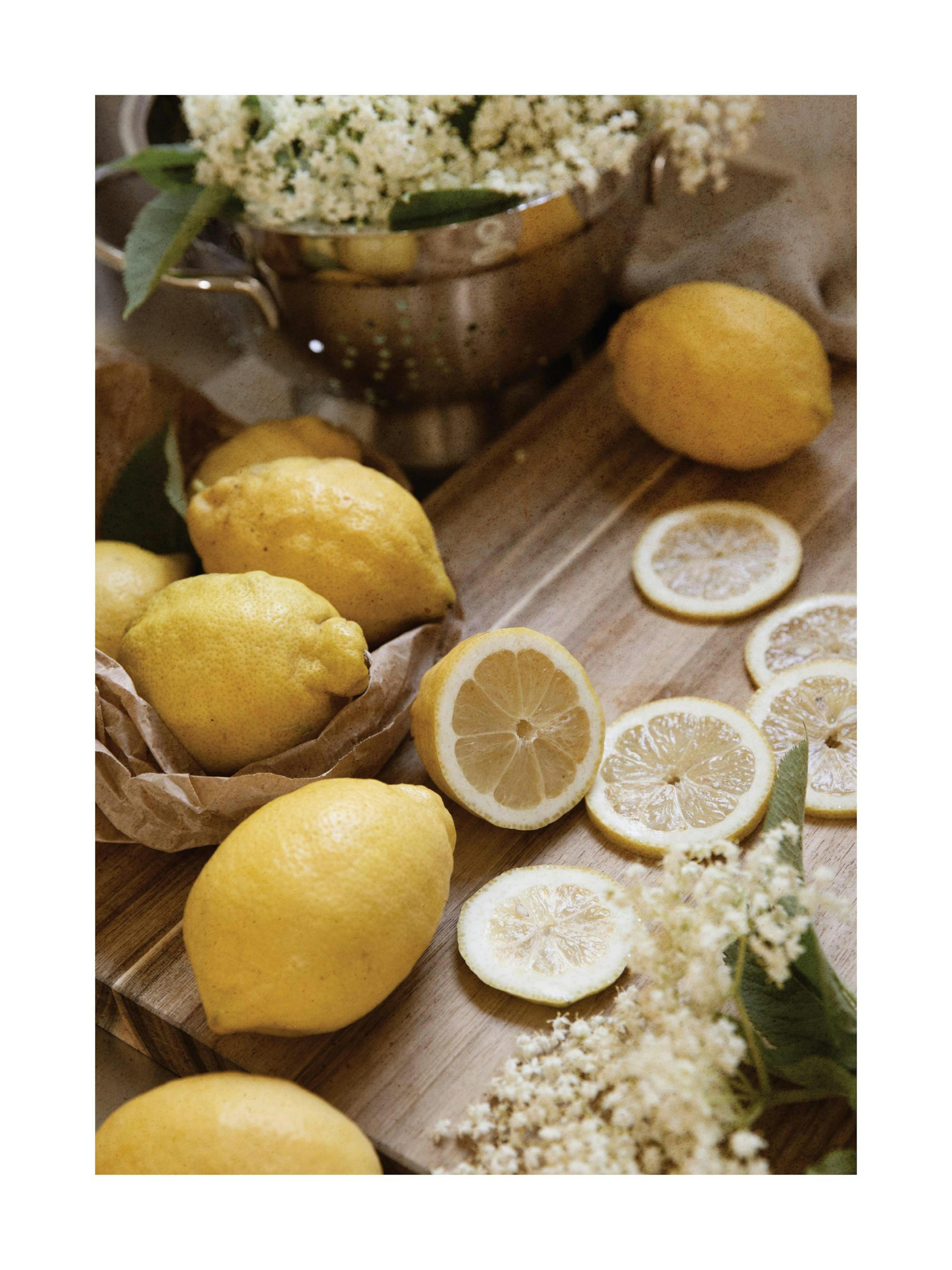 A poster featuring a still life of lemons and cut lemon slices on a wooden surface, with a colander of white elderflowers in the