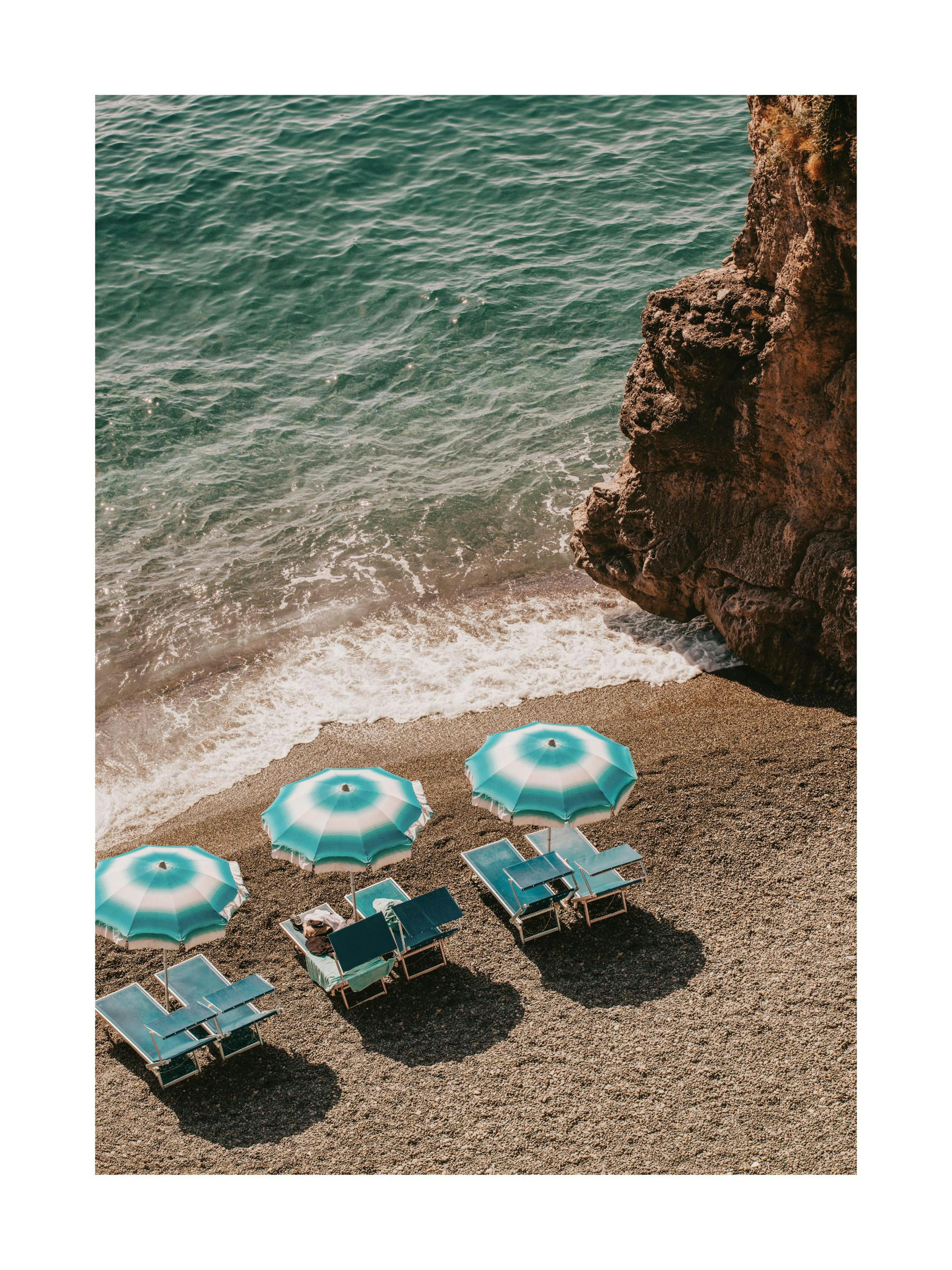 A poster featuring a beach with turquoise umbrellas and lounge chairs, next to ocean waves and a cliff, hanging above a table wi