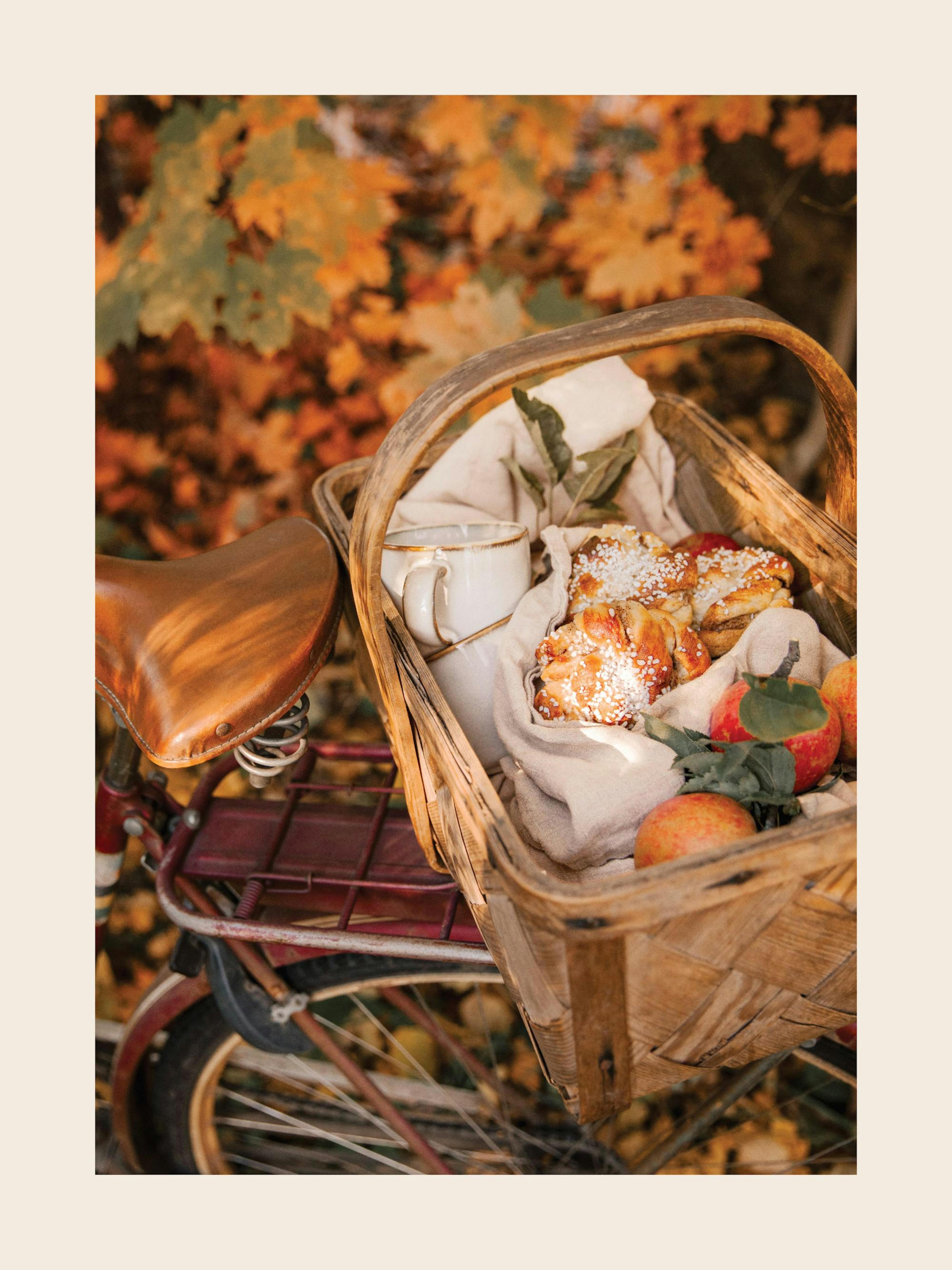 A poster featuring a rustic bicycle with a basket filled with baked goods, apples, and a mug, set against autumn leaves.