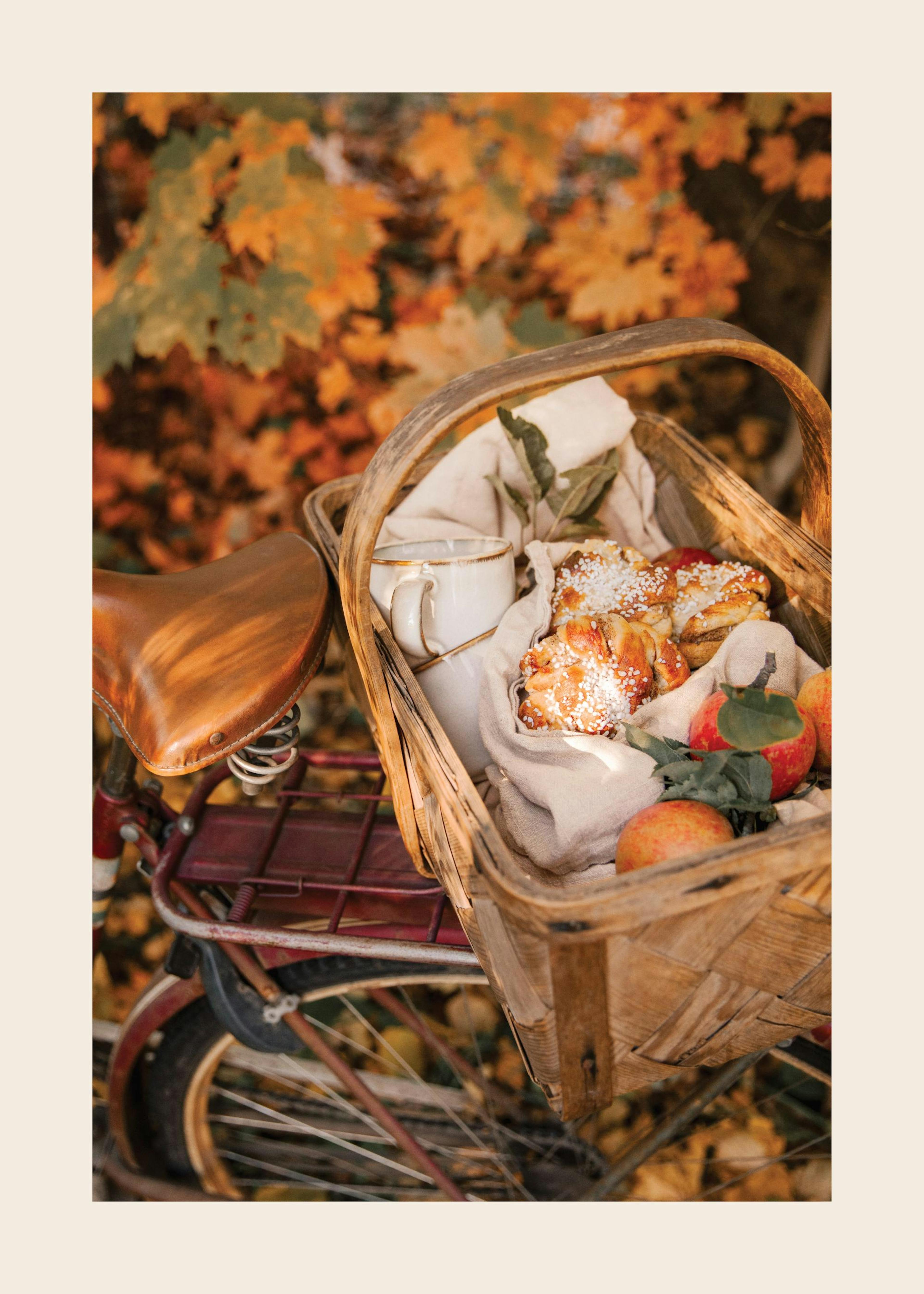 A poster featuring a rustic bicycle with a basket filled with baked goods, apples, and a mug, set against autumn leaves.