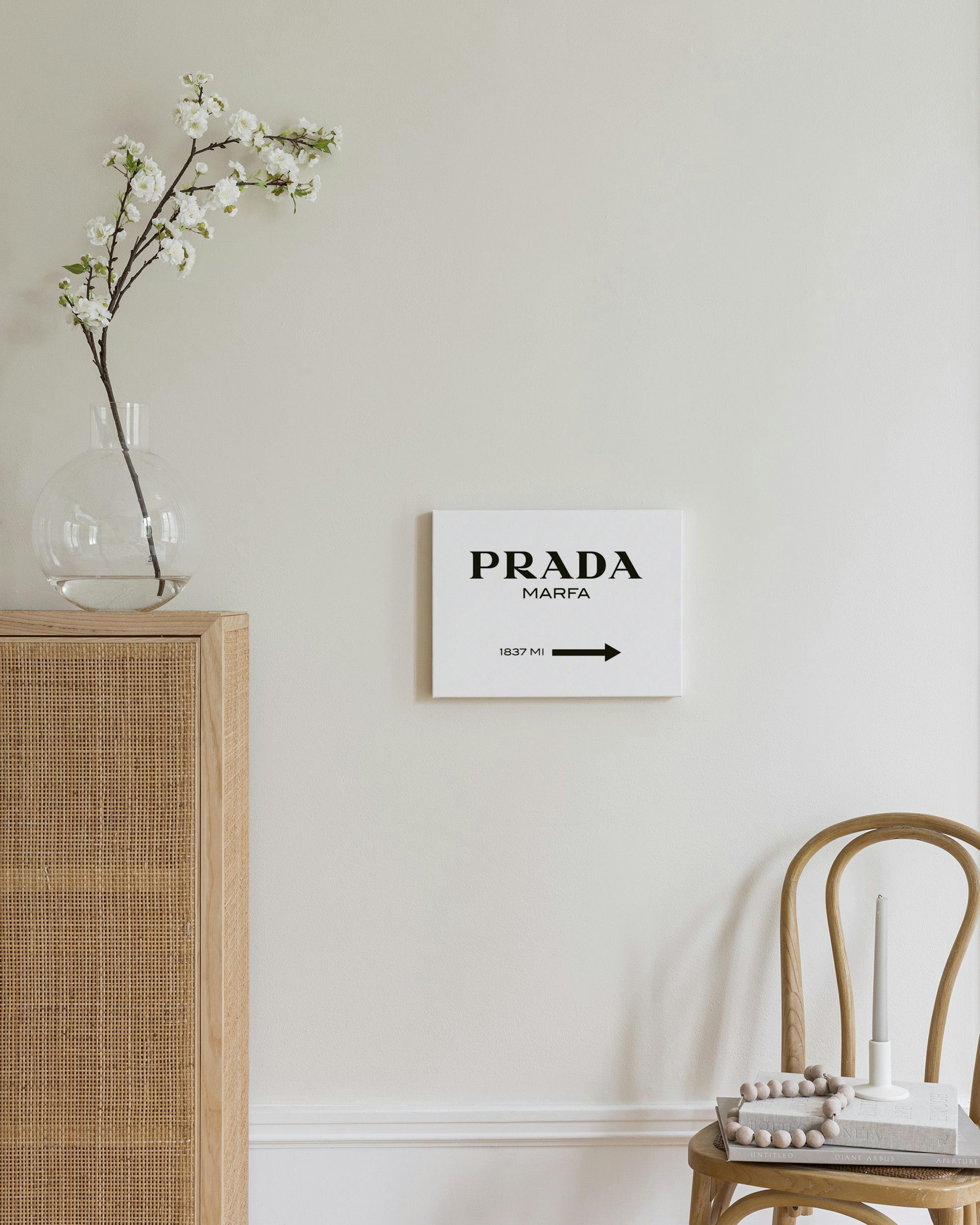 A white canvas with PRADA MARFA 1837 MI and an arrow, hanging above a wooden rattan dresser with a lamp and decor.