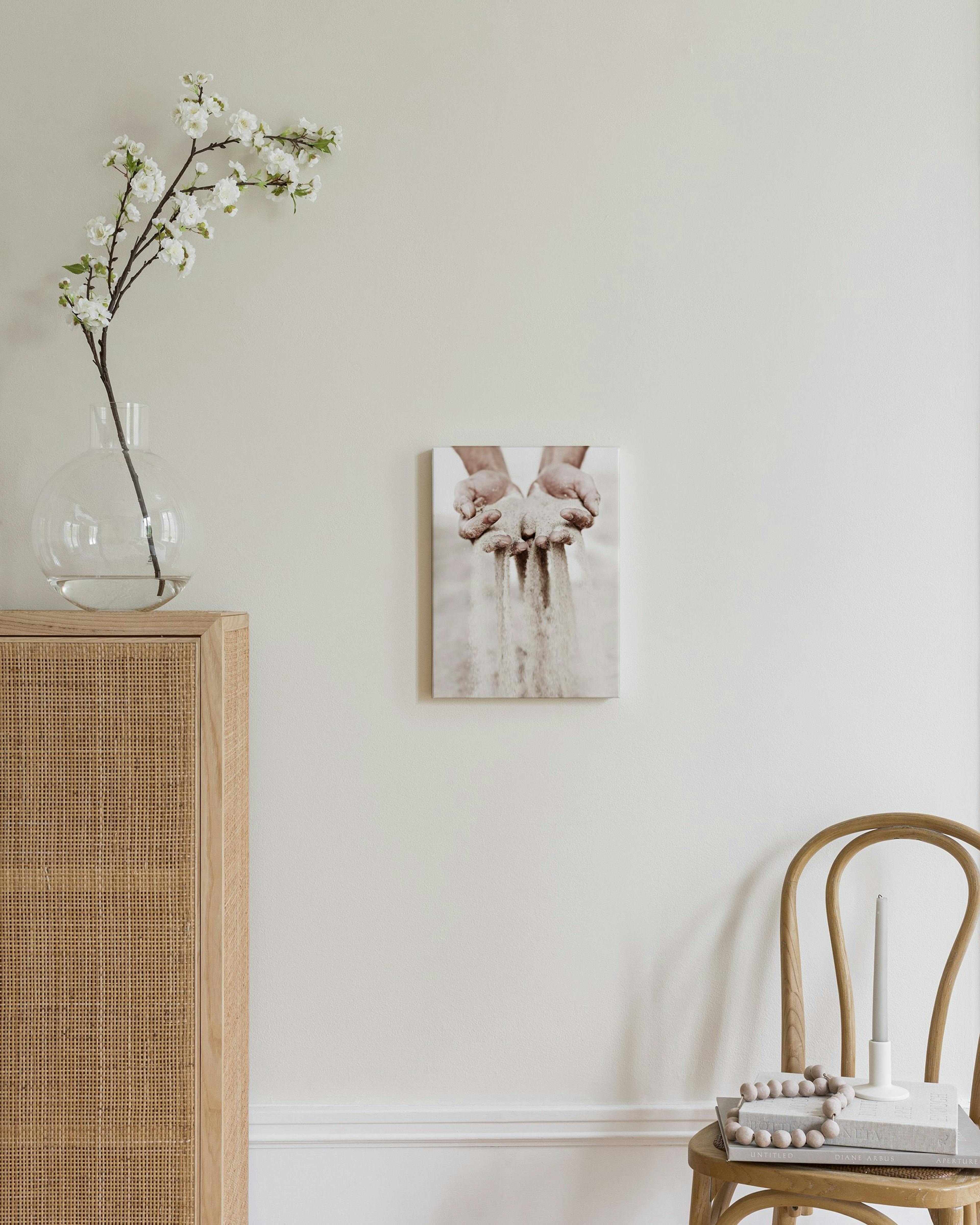 A canvas showing hands sifting sand, displayed in a light-colored room with a white vase on a pedestal and a textured ottoman.
