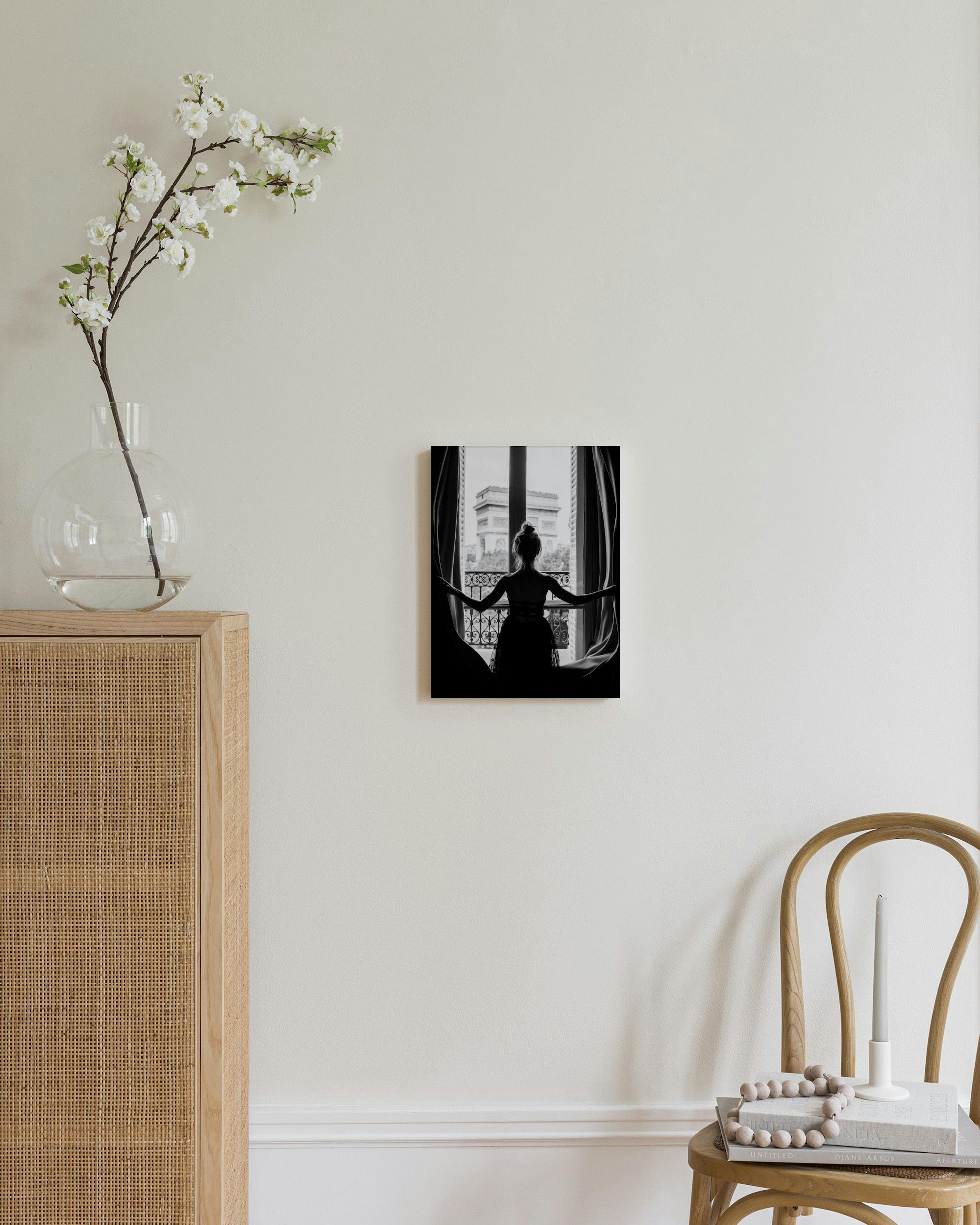 Black and white print of a woman opening curtains to reveal the Arc de Triomphe, displayed in a minimal room.