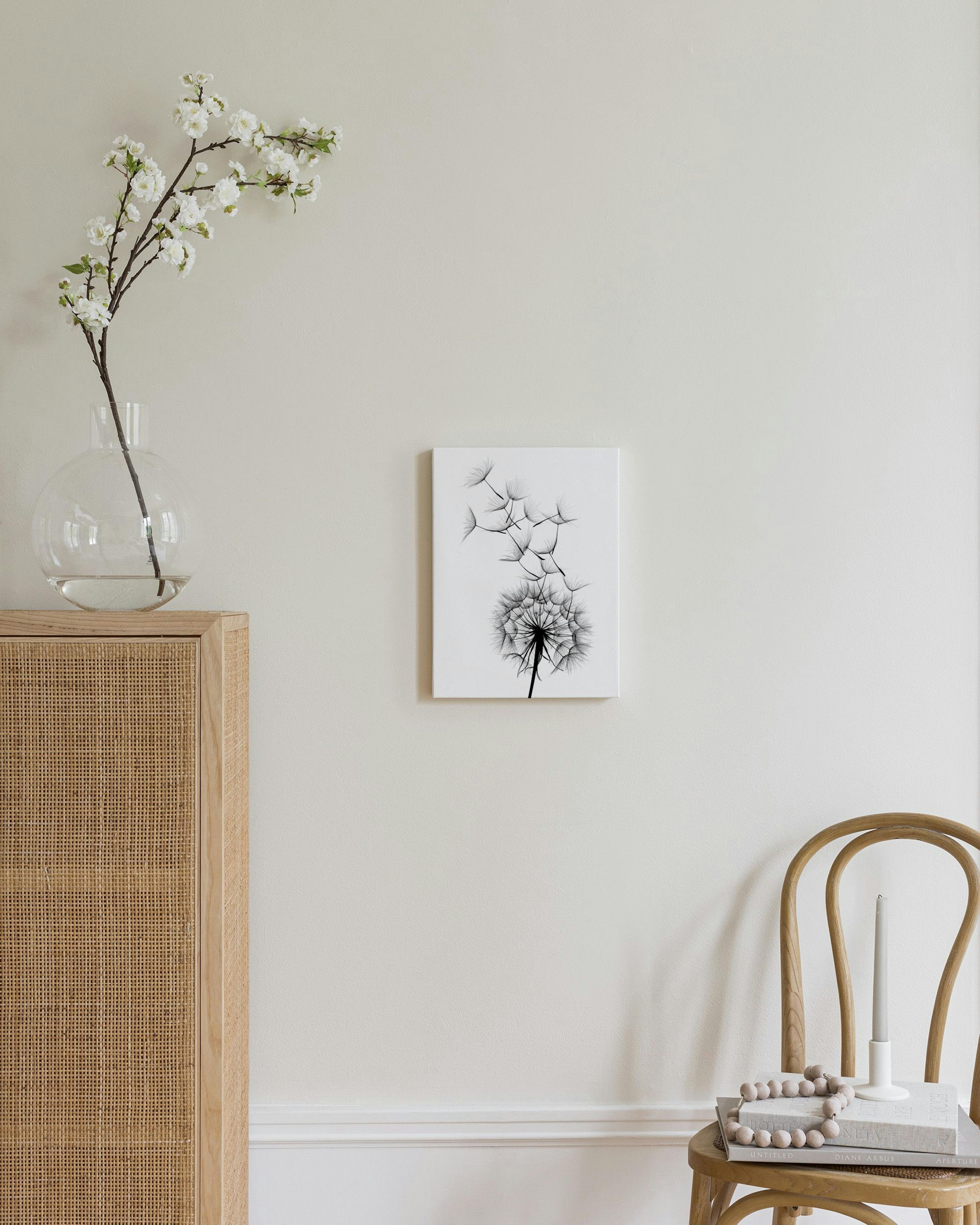 Canvas met zwart-wit paardenbloem en wegvliegende pluizen, hangend in een moderne, minimalistische kamer.
