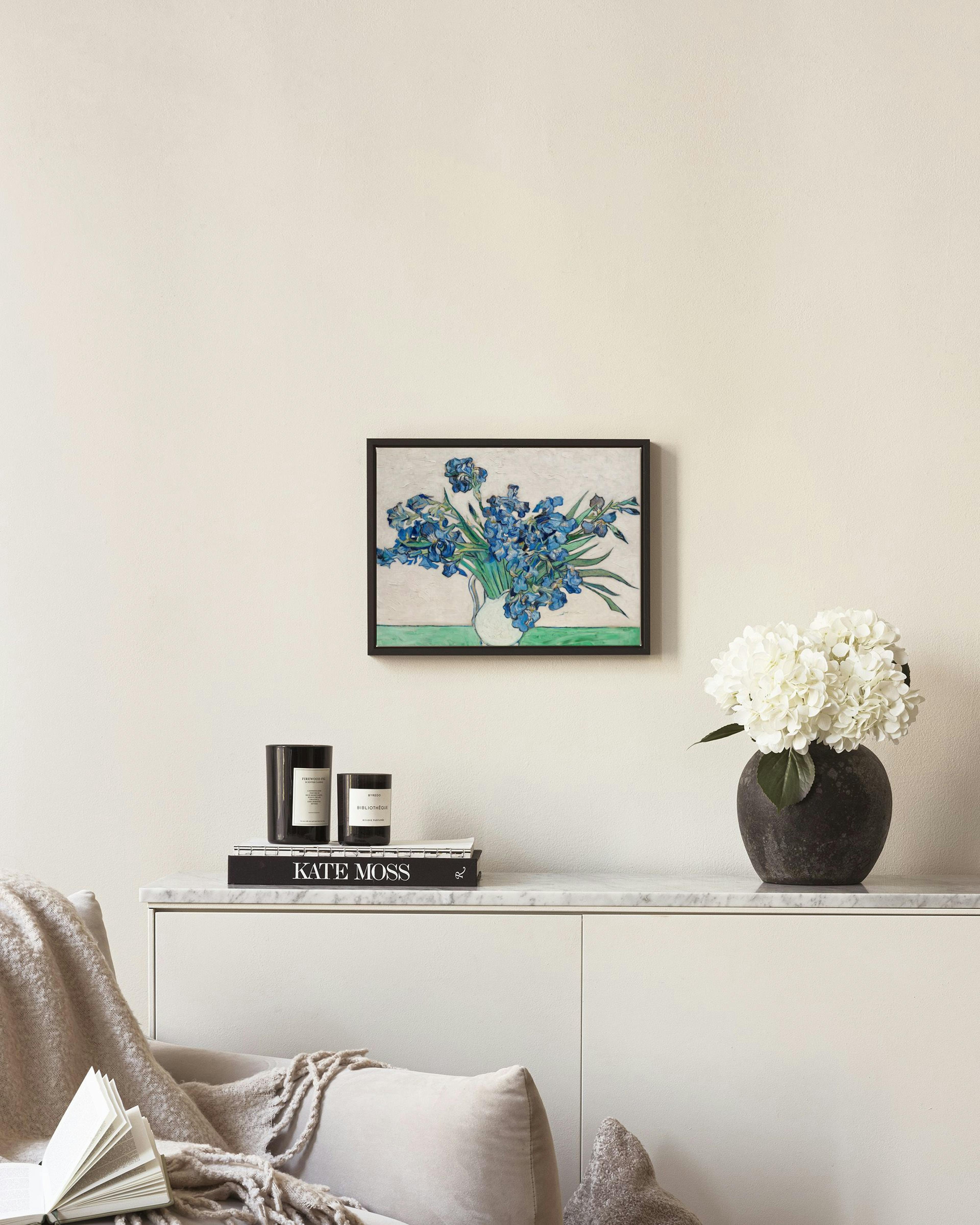 A canvas of blue irises in a white vase, hanging on a light wall above a wooden chair with decor.