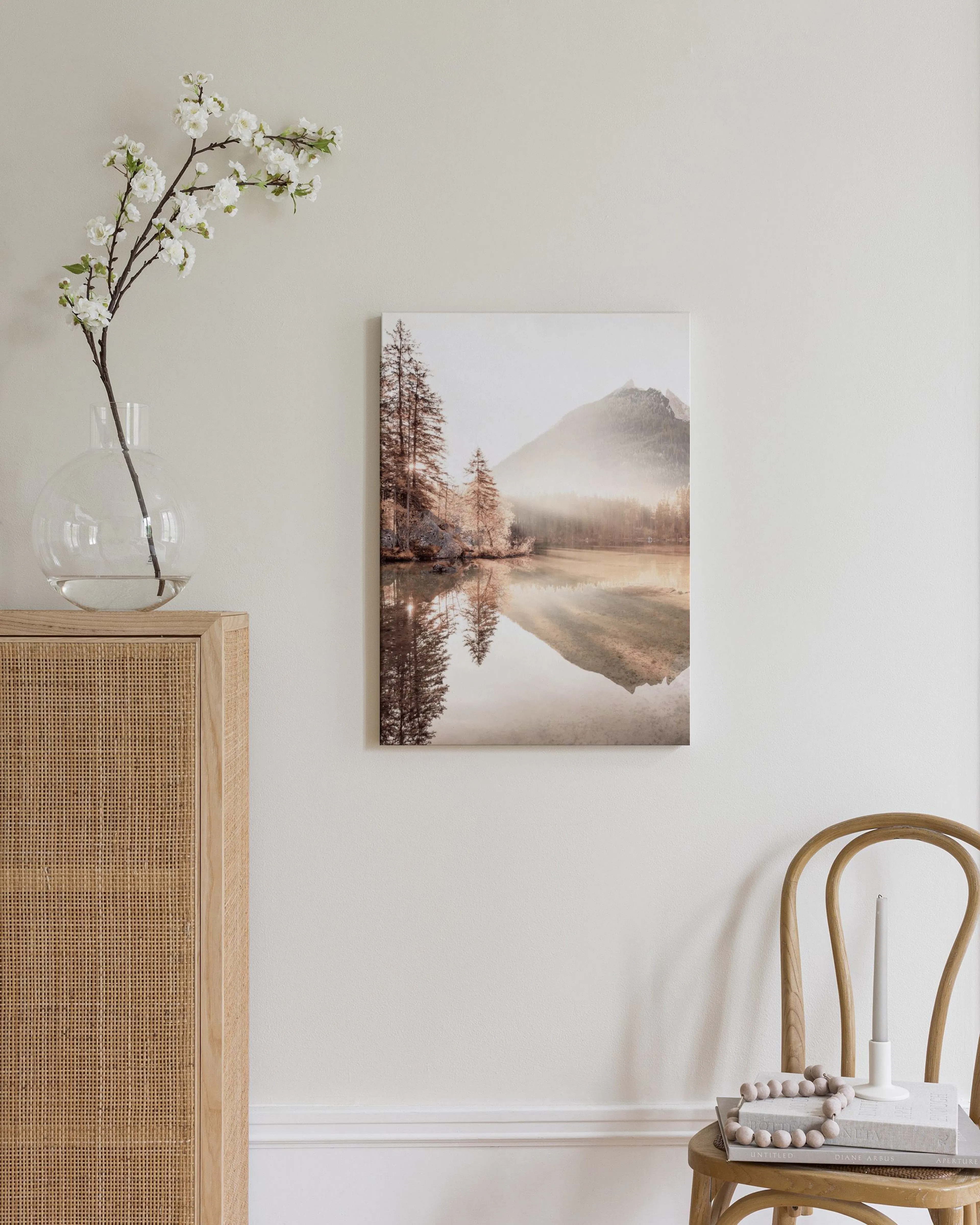 Mountain lake landscape with trees and reflections, hanging above a wooden rattan cabinet with a lamp and books.