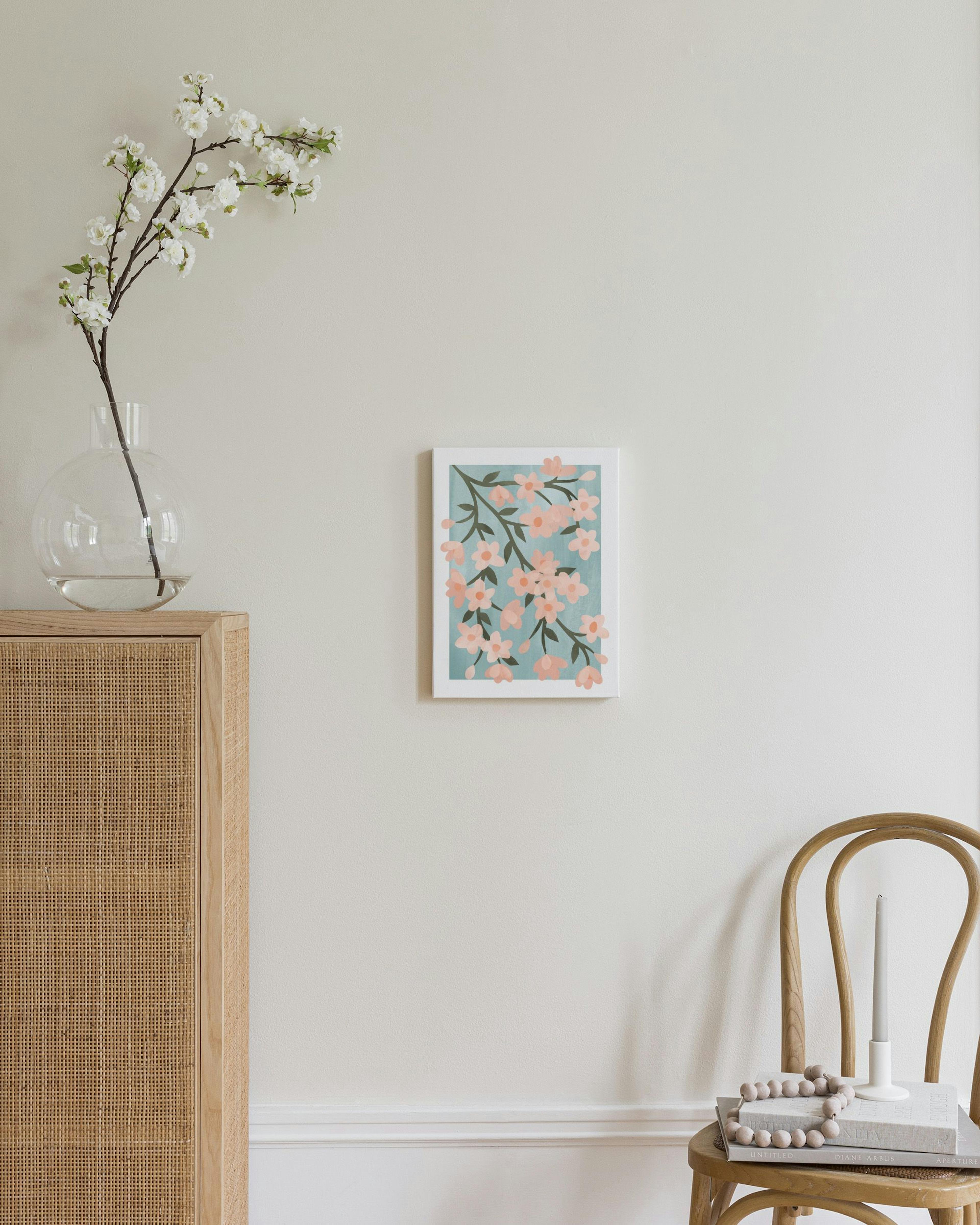 Lienzo de ramas floreciendo con flores rosadas y hojas verdes sobre fondo azul claro, exhibido sobre un mueble de madera.