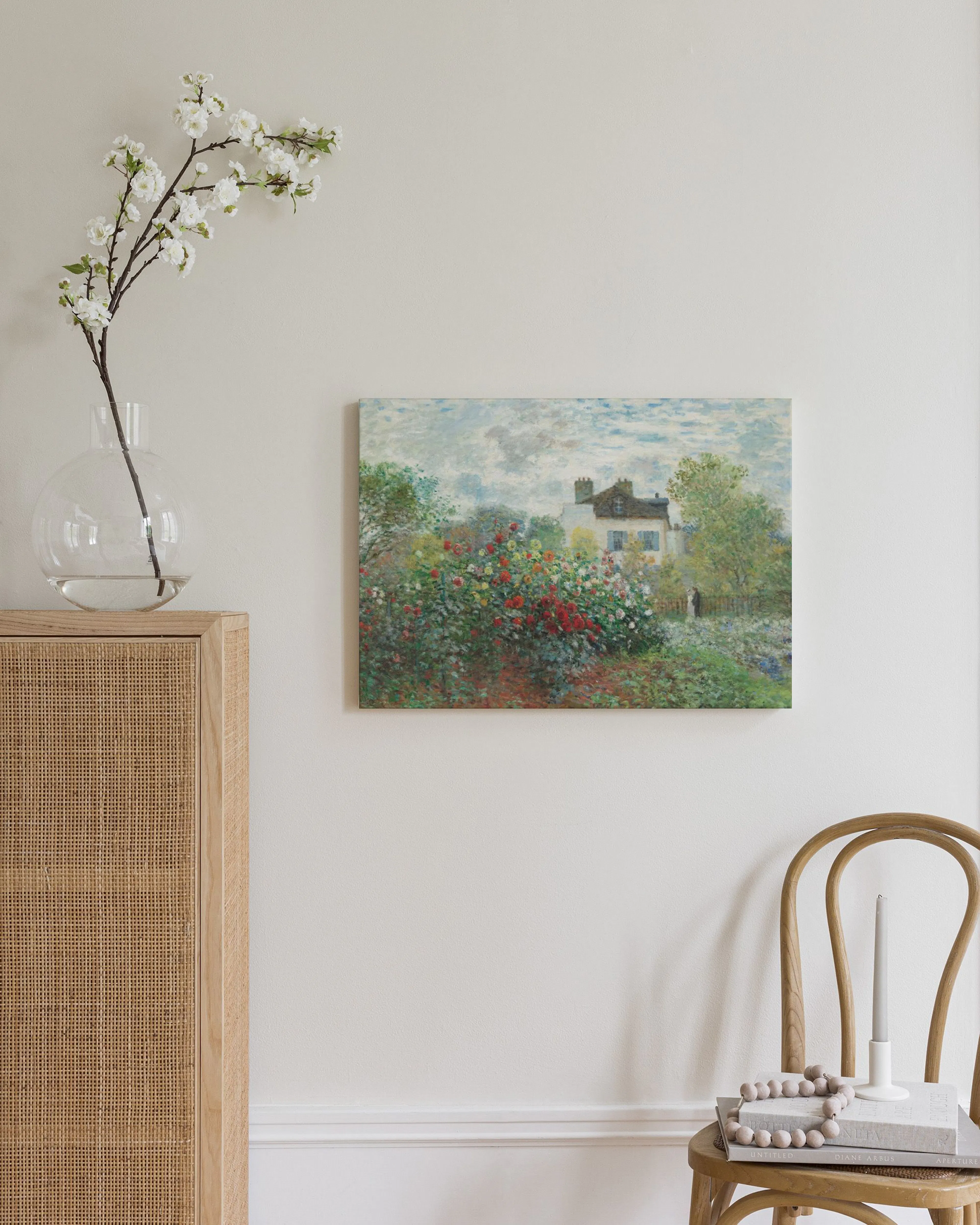 Impressionist painting of a house surrounded by a vibrant flower garden, hanging on a white wall above a wooden chair with books